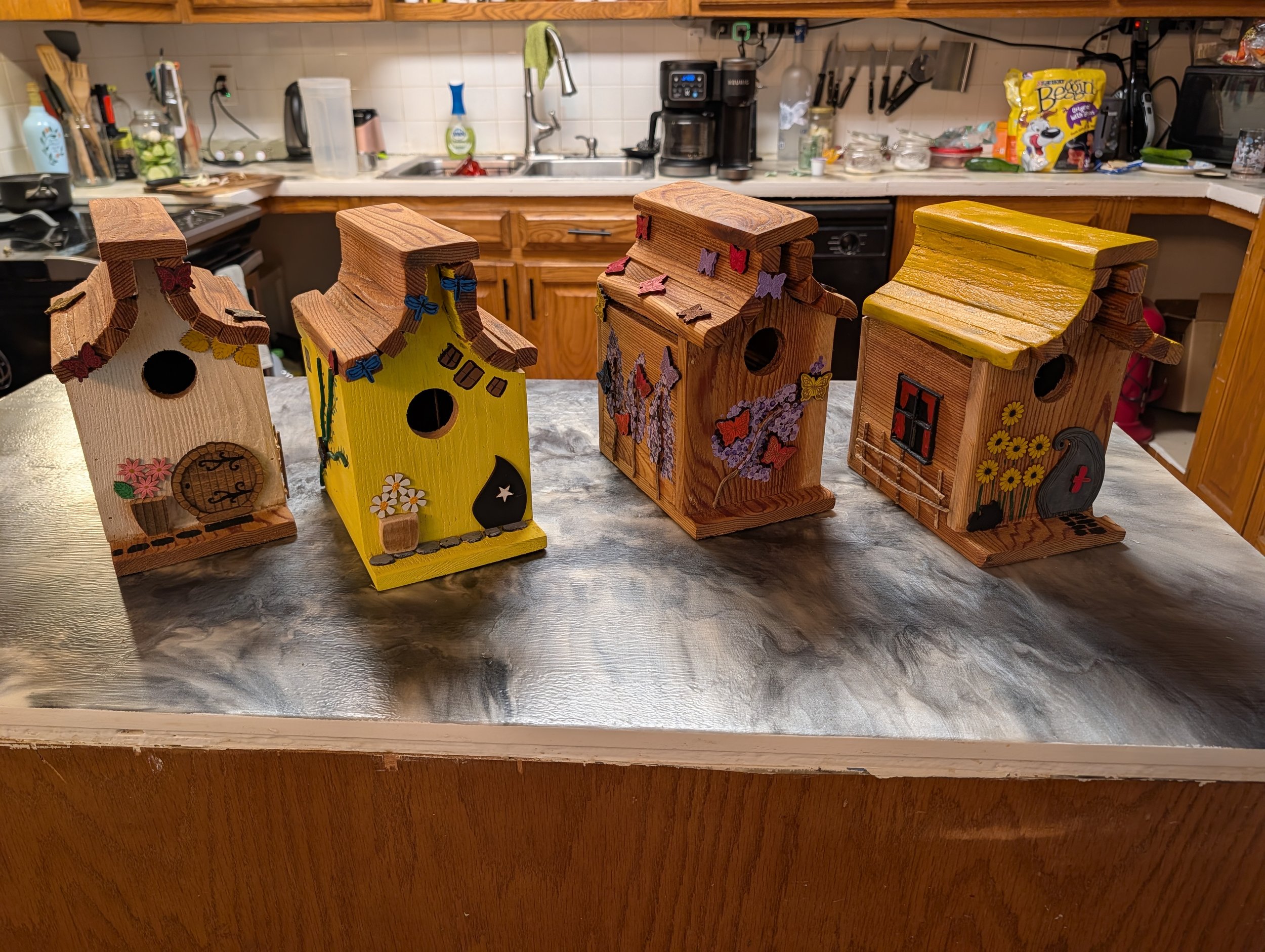 Four colorful wooden birdhouses with various decorative elements placed on a kitchen countertop.