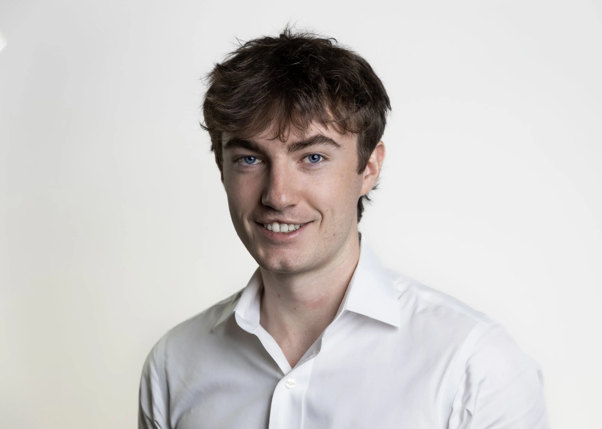 A young man with brown hair and blue eyes, wearing a white dress shirt, smiling in front of a plain light-colored background.