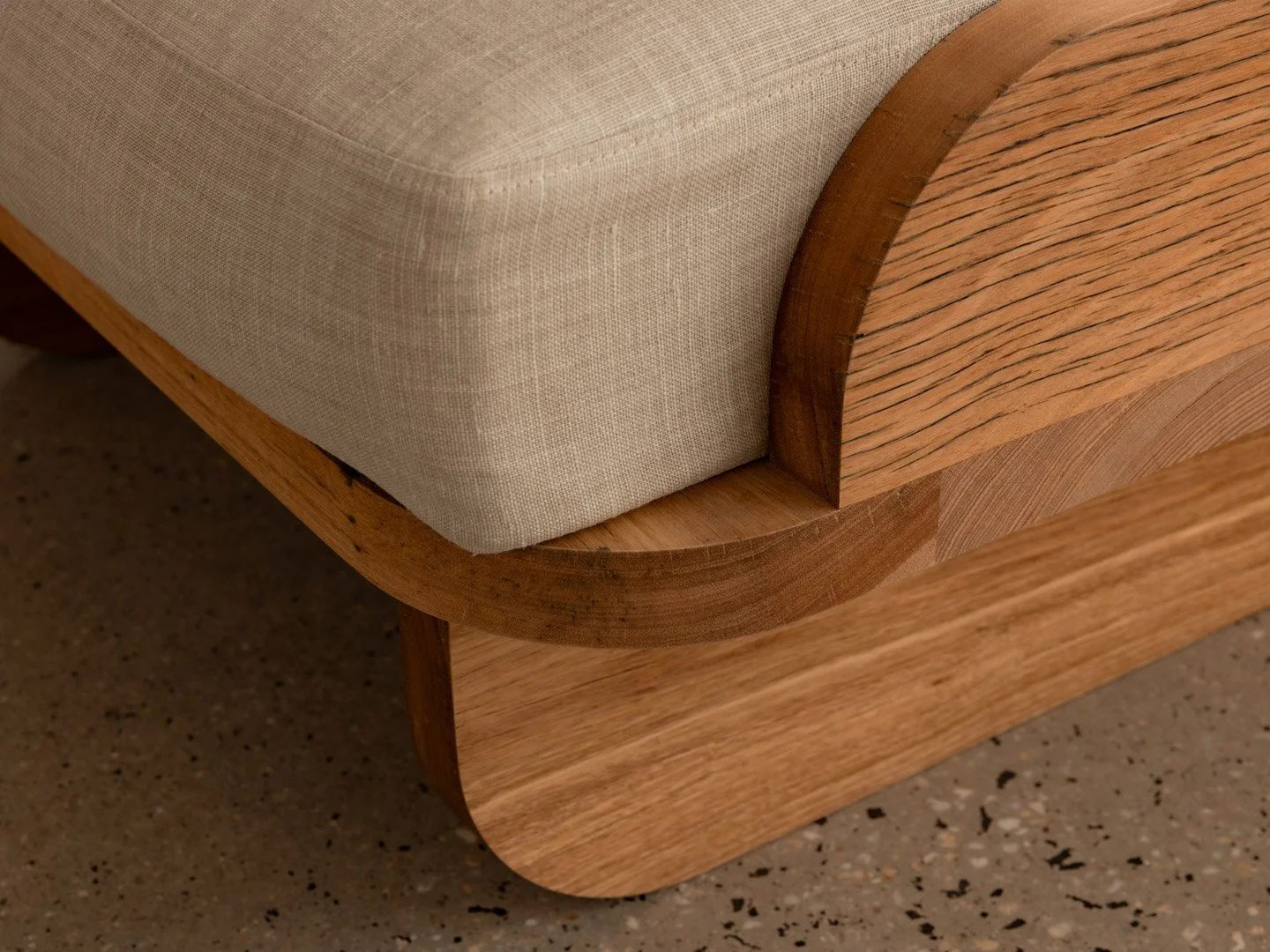 Close-up of a wooden bed frame with a rounded corner, featuring a beige upholstered mattress or cushion, on a speckled floor.