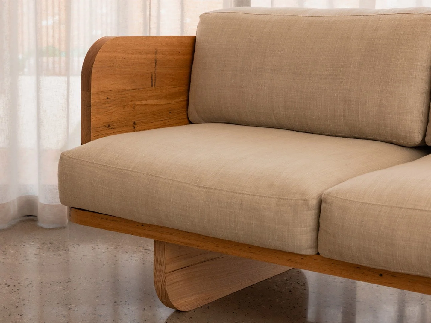 Close-up of a beige upholstered sofa with wooden armrests and support, positioned near a window with sheer curtains.