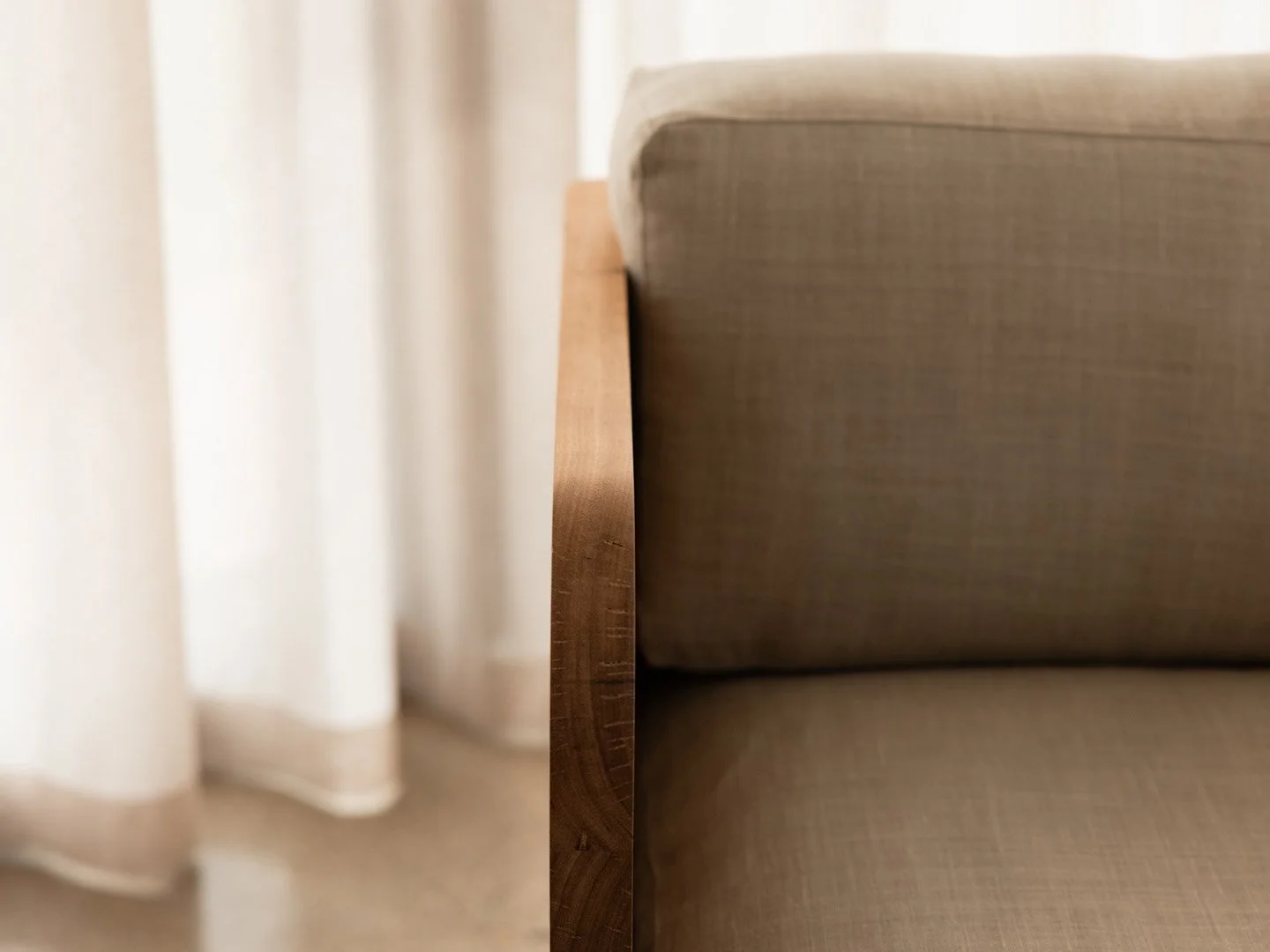 Close-up of a beige armchair with a wooden side panel in a room with cream-colored curtains.
