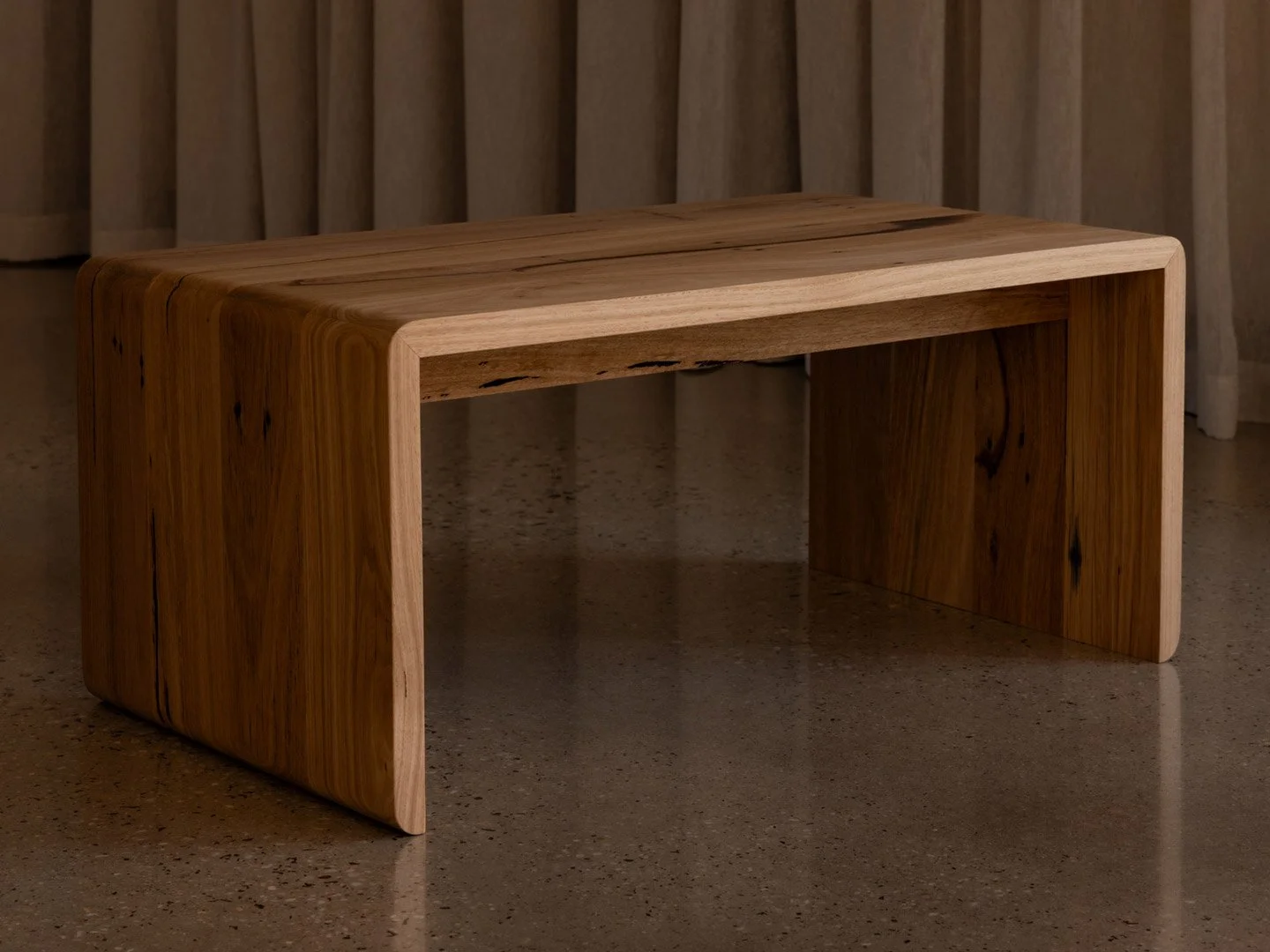 A modern wooden side table with a minimalist design, smooth curves, and natural wood finish, placed on a speckled concrete floor with a background of vertical wooden slats.