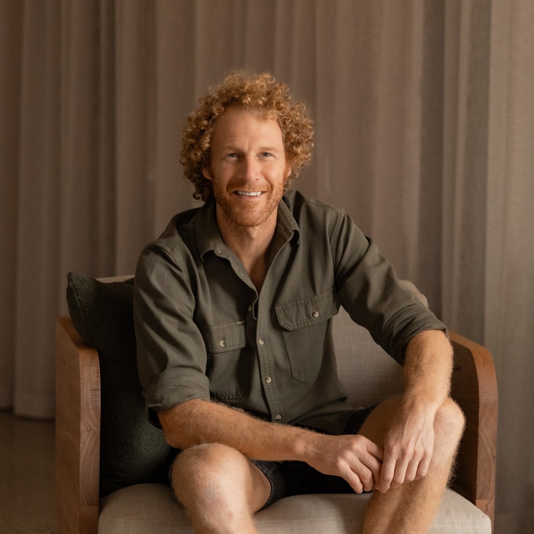 A man with curly hair and a beard sitting on a chair in a room with beige curtains, smiling at the camera.