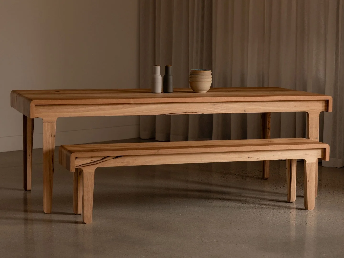 A wooden dining table with matching bench, set against a neutral background with curtains, with three small ceramic bottles and a stack of bowls on top.
