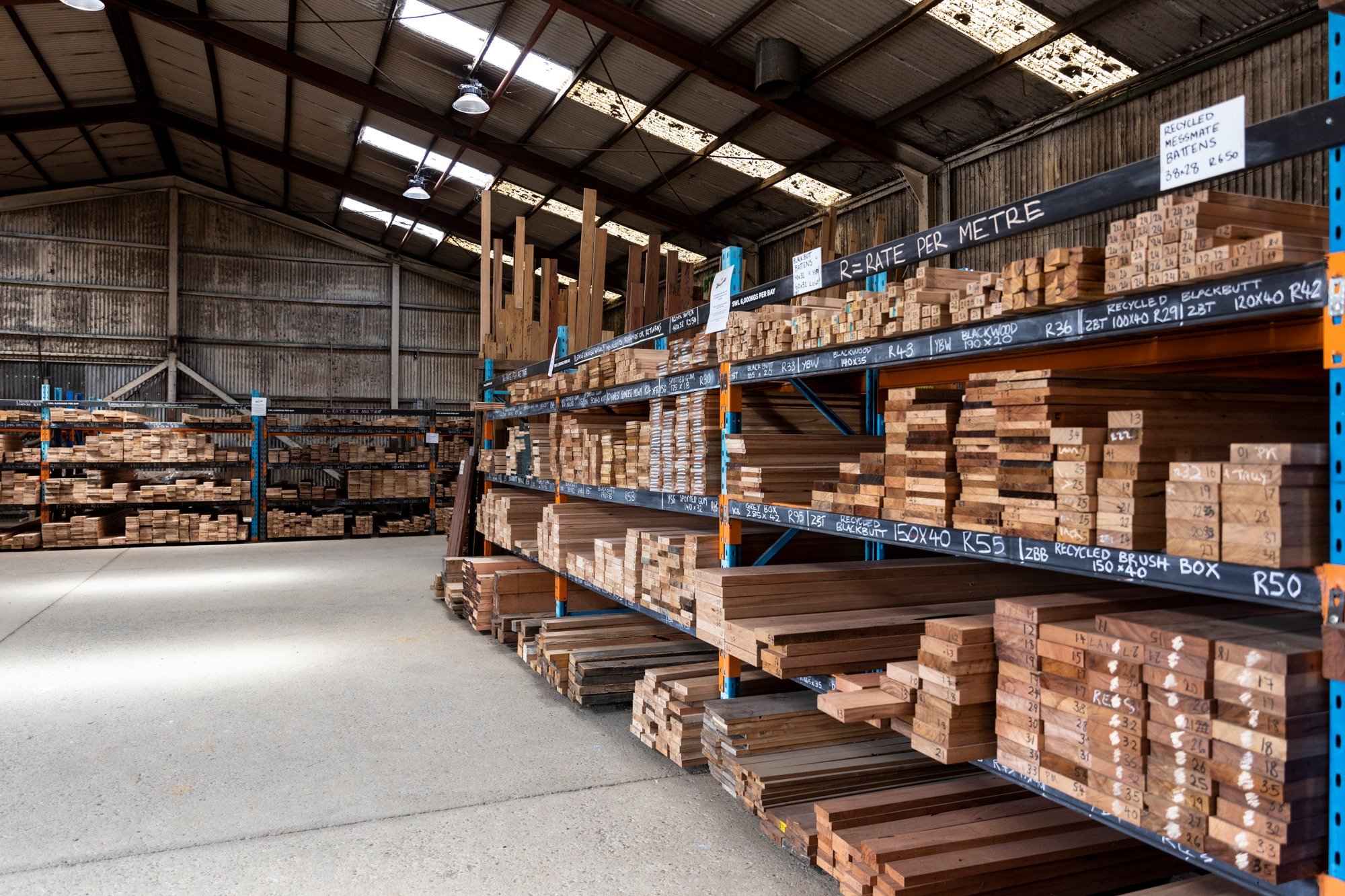 Shelves of wooden planks and timber inside a warehouse with metal roof panels and skylights.