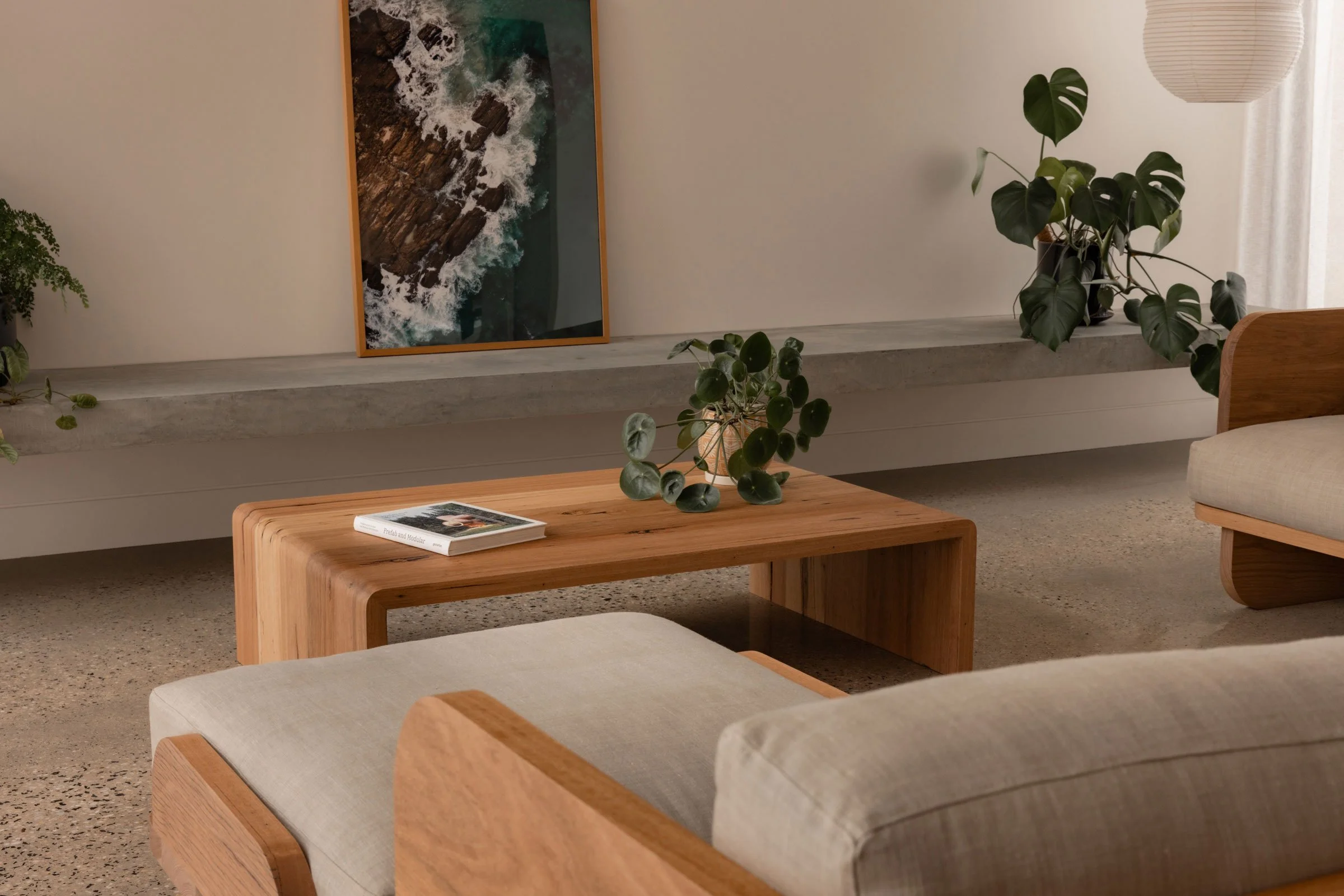 Living room with a wooden coffee table, potted plants, a framed aerial photo of ocean waves on a ledge, a part of a sofa, and a hanging paper lantern.