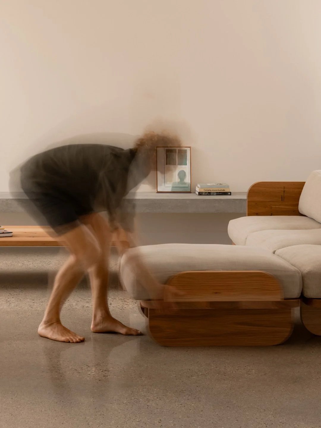 A person bending over with a motion-blurred form, in a living room with a beige sofa, wooden armrest, a bench, a picture frame, and a stack of books on a wall-mounted shelf.