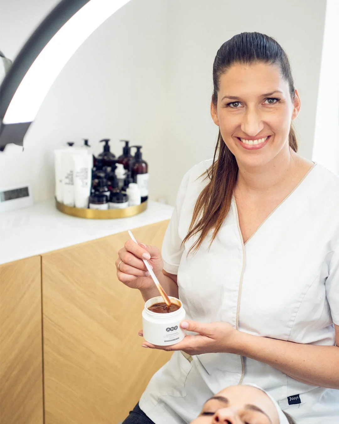 Eine Frau, wahrscheinlich Kosmetikerin, die in einem Behandlungsraum kosmetische Produkte aus einem weißen Behälter entnimmt, während sie lächelt.