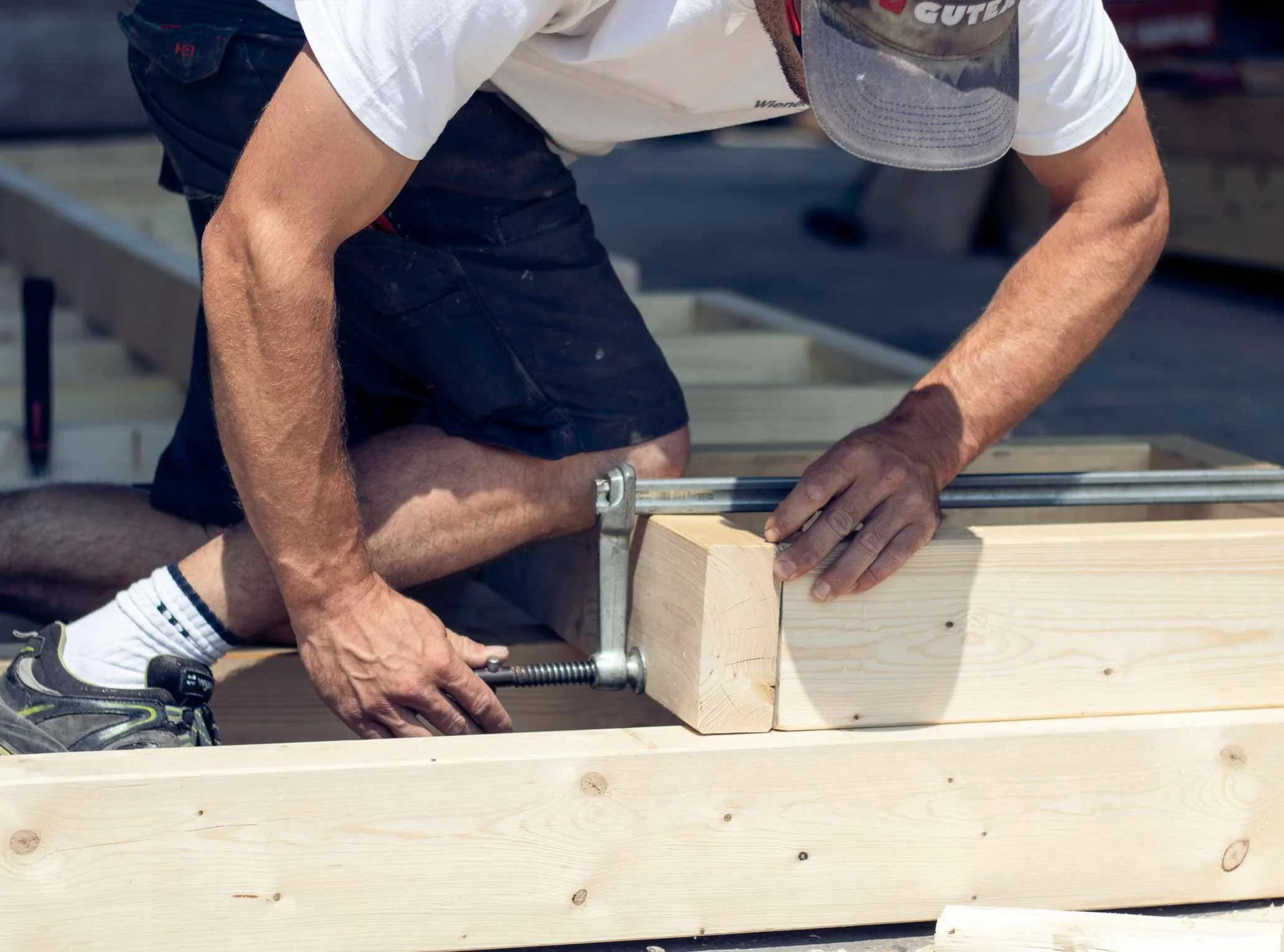 Ein Mann arbeitet mit einer Zwinge an einem Holzstück, trägt einen Cap, ein weißes T-Shirt und schwarze Shorts, auf einem Baugrundstück.