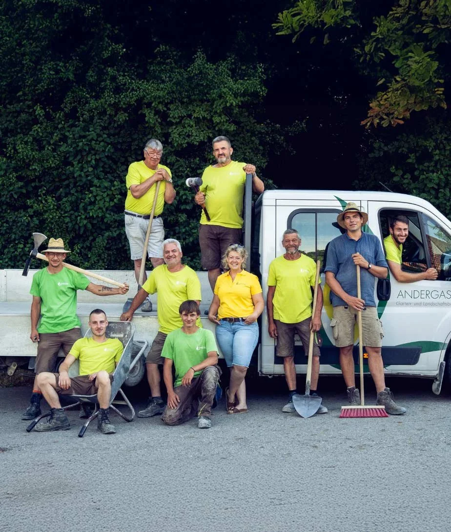 Gruppen von Menschen bei einer Gemeinschaftsarbeit im Freien, einige tragen Werkzeuge, stehen und sitzen vor einem weißen Fahrzeug, umgeben von grünen Bäumen.