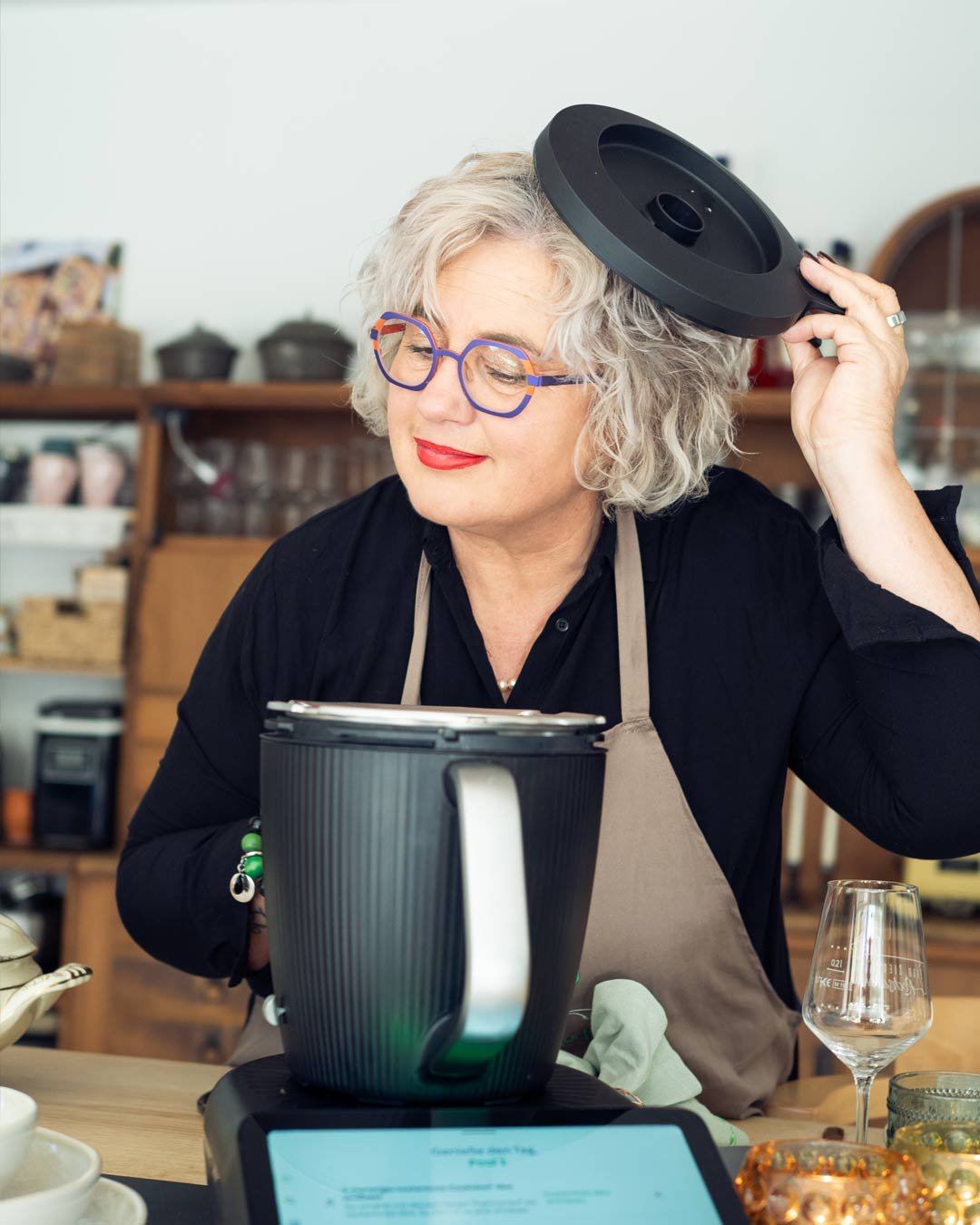 Ältere Frau beim Kochen in einer Küche, trägt eine Kochschürze, hat graues, lockiges Haar und trägt bunte Brillen, hält eine runde schwarze KitchenAid-Mixer-Außenhaube über dem Kopf.