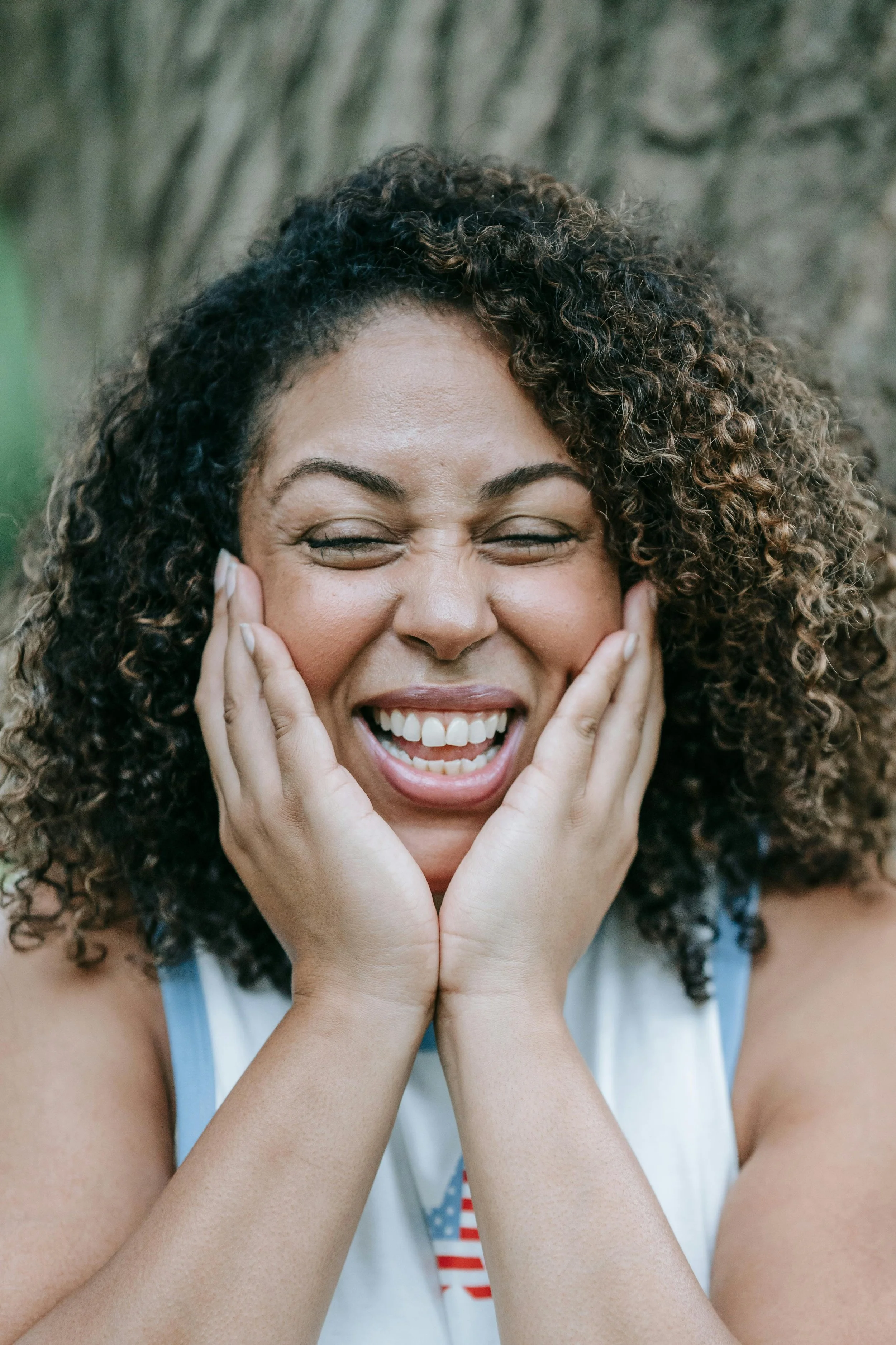 A woman with curly hair is smiling joyfully with her eyes closed, her hands gently holding her face.