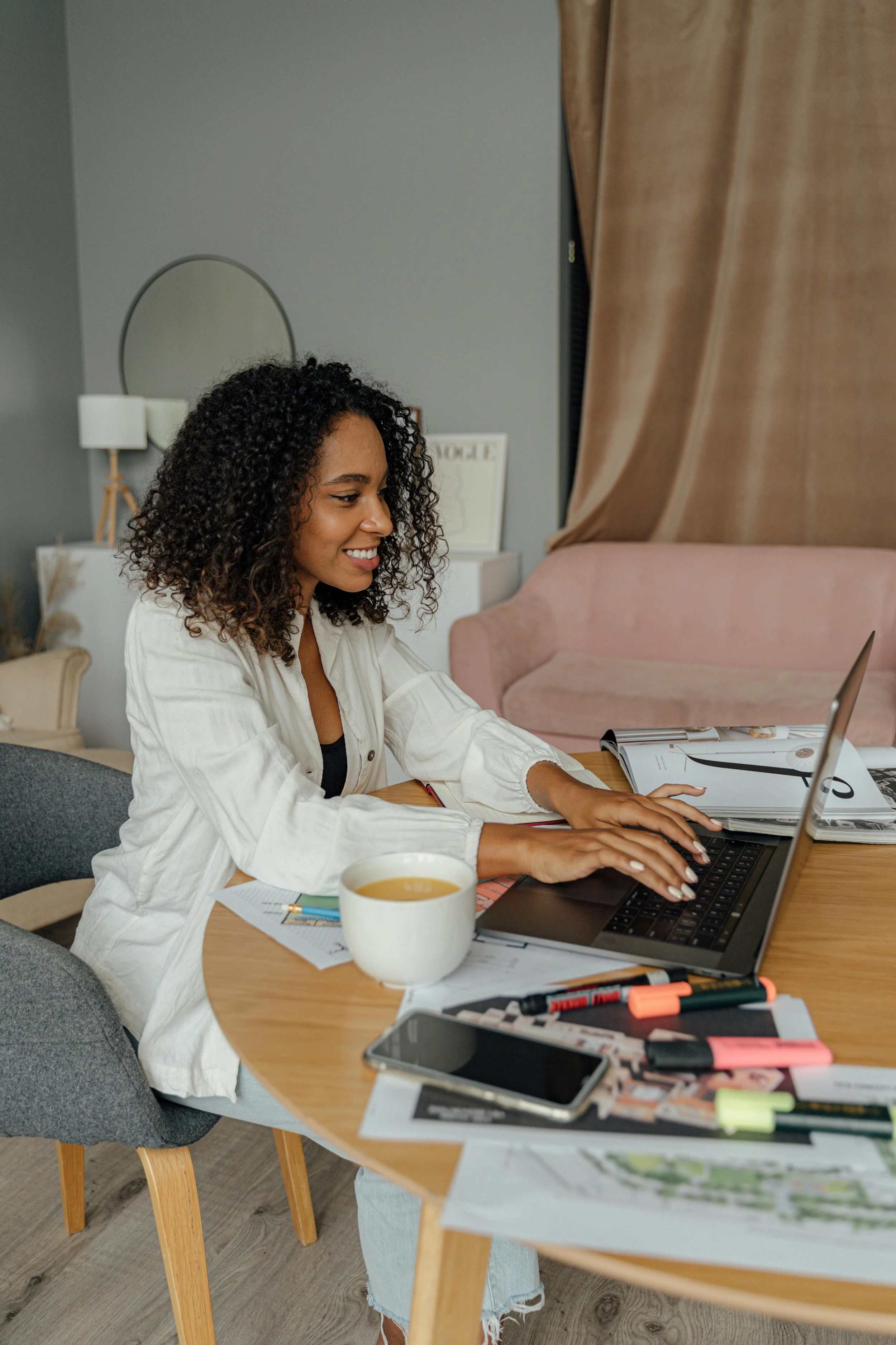 A woman with curly hair, busy entrepreneur, working on a laptop at a cluttered wooden table with notebooks, pens, highlighters, a smartphone, and a mug of tea in a cozy, modern living room.
