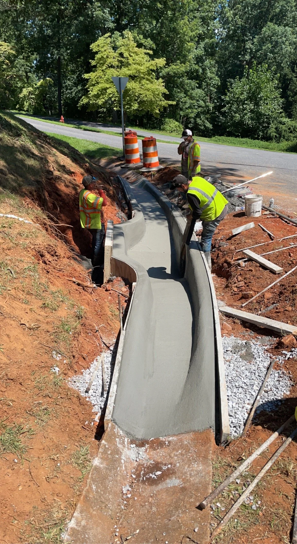 Construction workers building a concrete sidewalk beside a road surrounded by trees, with orange traffic barrels and a yield sign nearby.