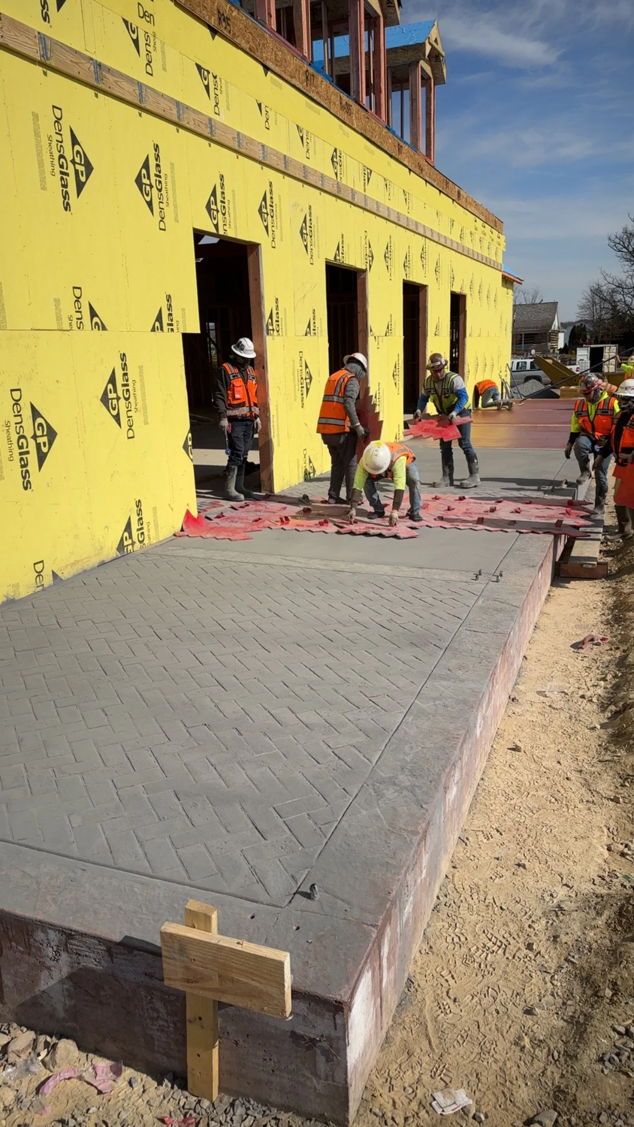 Construction workers pouring and finishing concrete on a sidewalk in front of a building under construction, with yellow insulation board on the building's exterior walls.