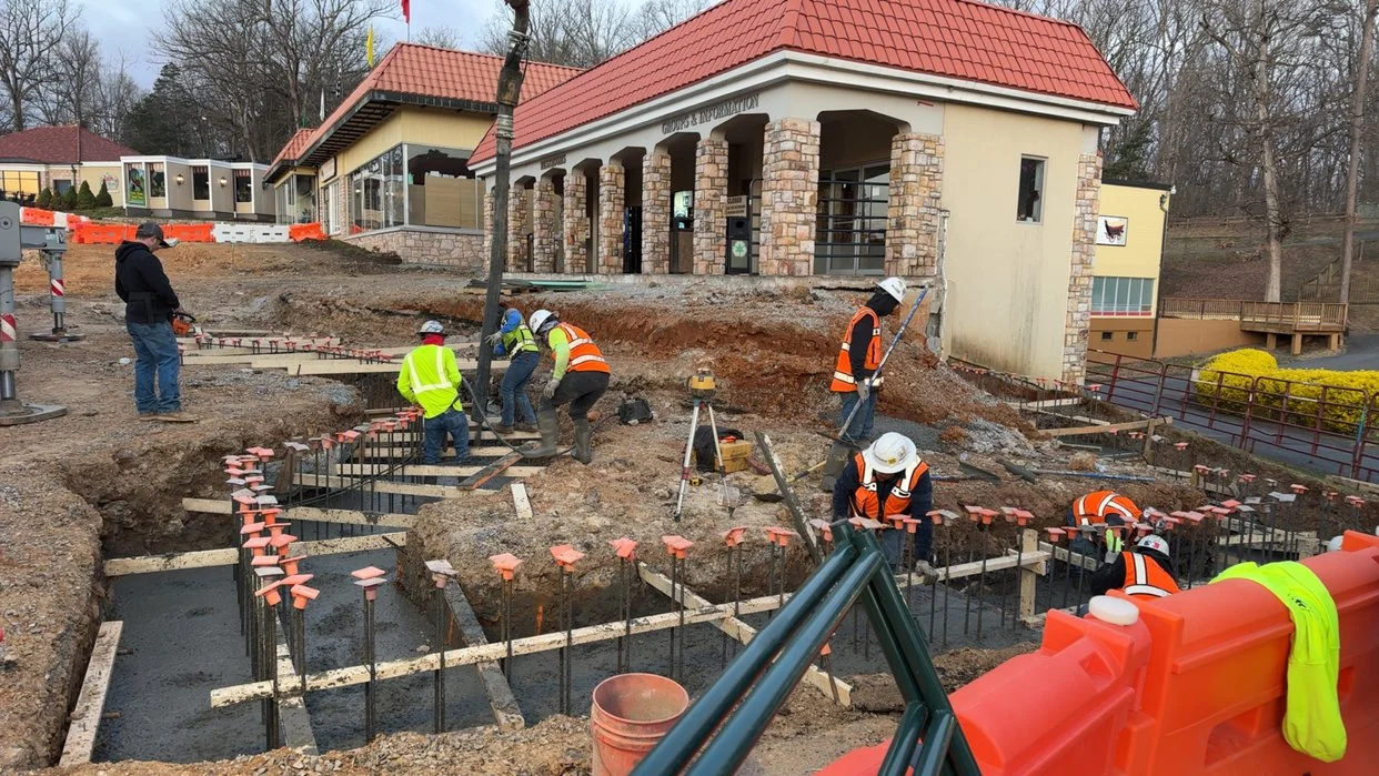 Construction workers in safety vests and helmets working on footings at a building site with a partly built structure in the background.