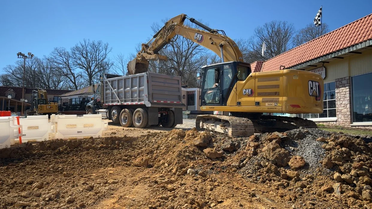 A construction site with a yellow CAT excavator loading dirt into a dump truck. The background shows a building with a red tile roof and leafless trees.