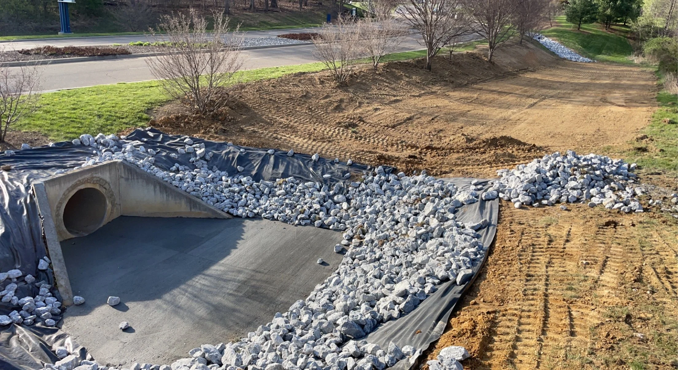 Construction site with a concrete drainage pipe, gravel, and dirt, with trees and grass in the background.