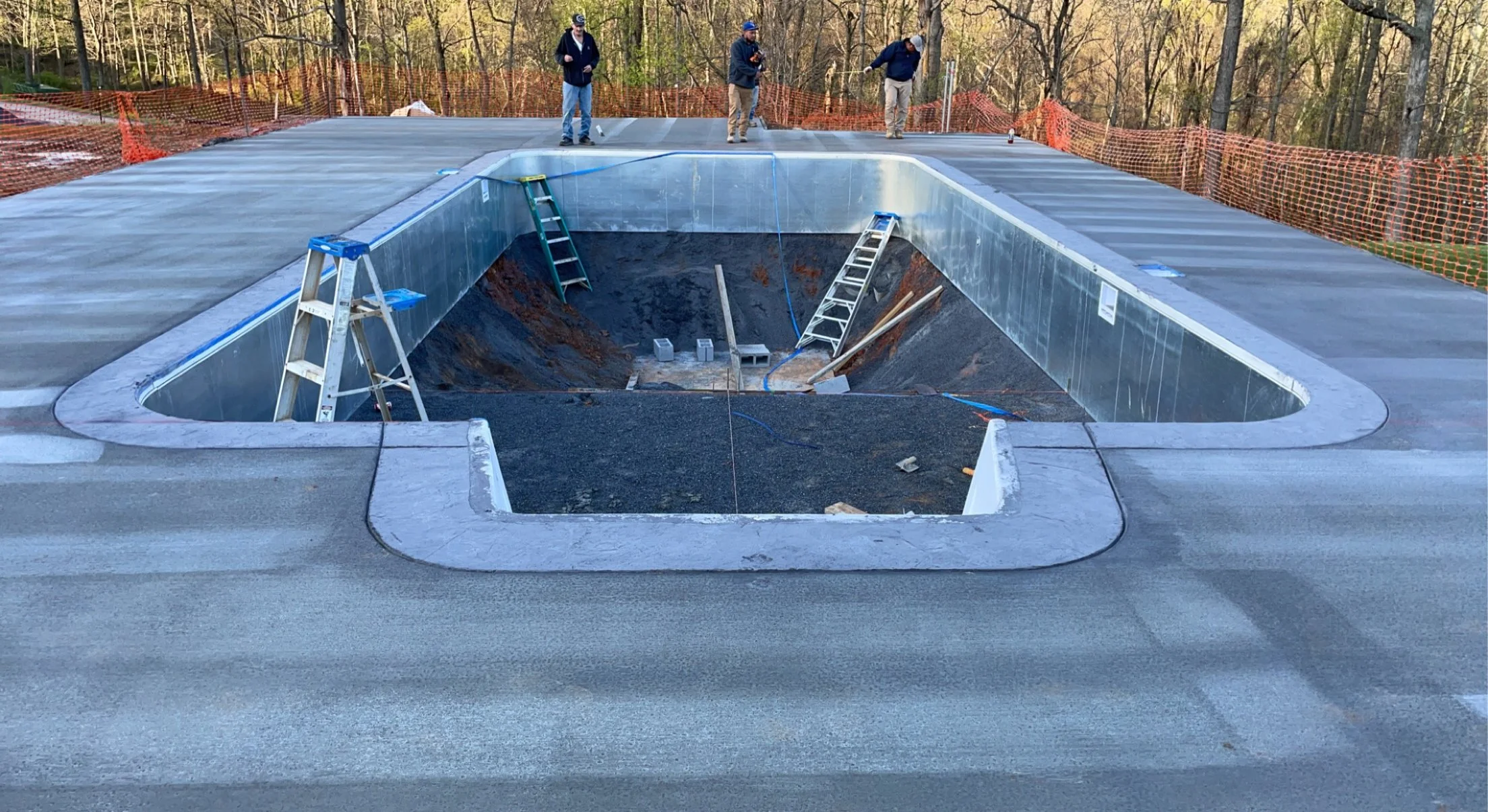 Construction workers work on the unfinished roof of a building with a large square skylight opening, surrounded by orange safety fencing, with trees in the background.