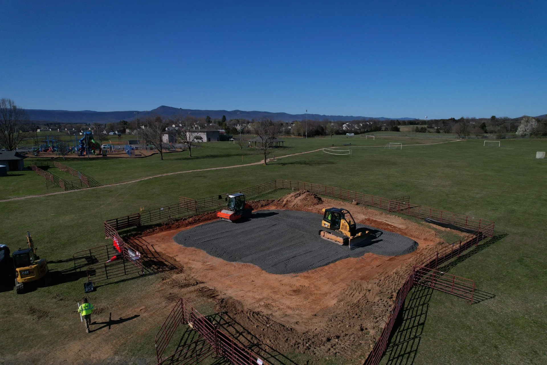 Construction site in a park with two excavators working on preparing the ground for a new field or playground. Fencing surrounds the construction area, and a worker in a yellow vest is standing nearby. In the background, there is a playground with sl