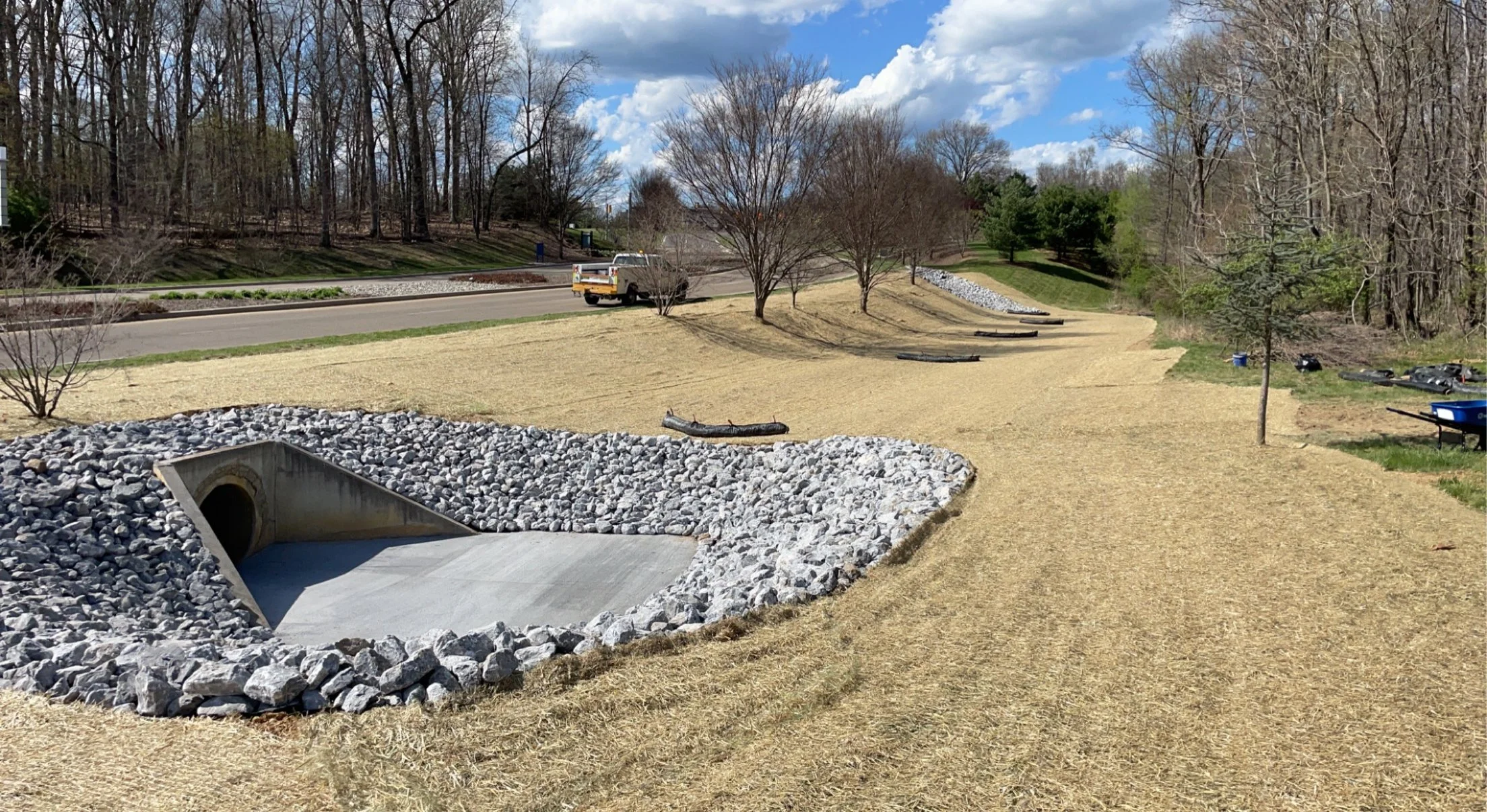 A landscaped park with a pathway, young trees, a water drainage culvert surrounded by rocks, and a pickup truck driving on the road in the background under a partly cloudy sky.