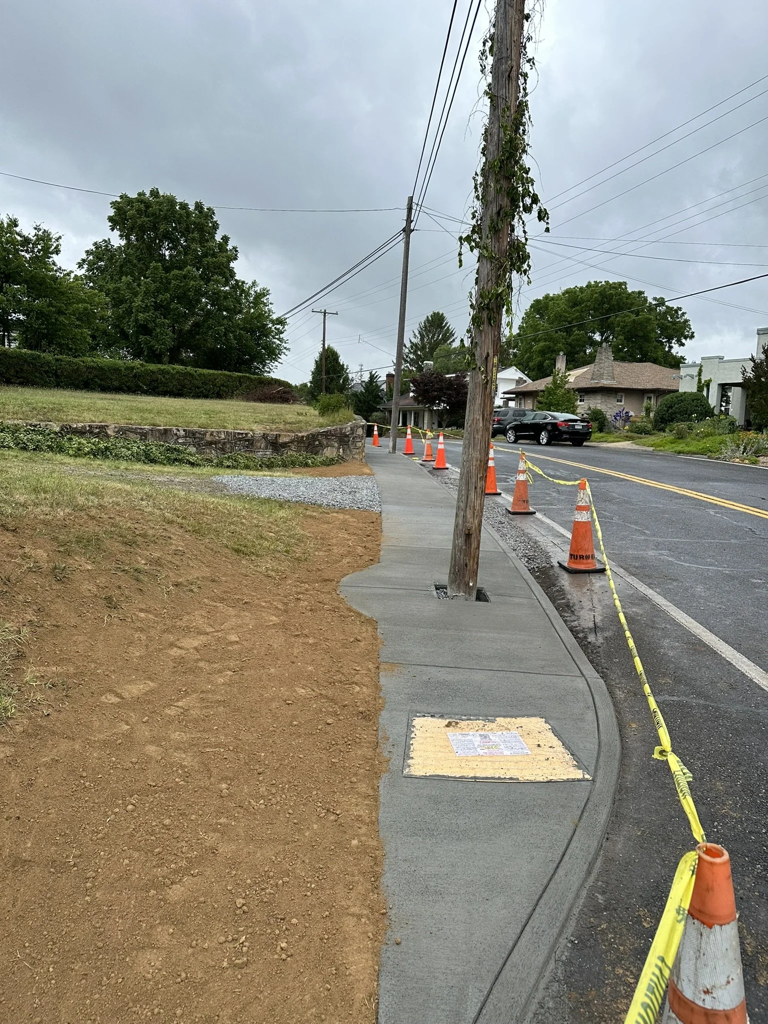 Newly poured concrete sidewalk with sidewalk access cover, a wooden utility pole, and orange construction cones and caution tape along the street under a cloudy sky.