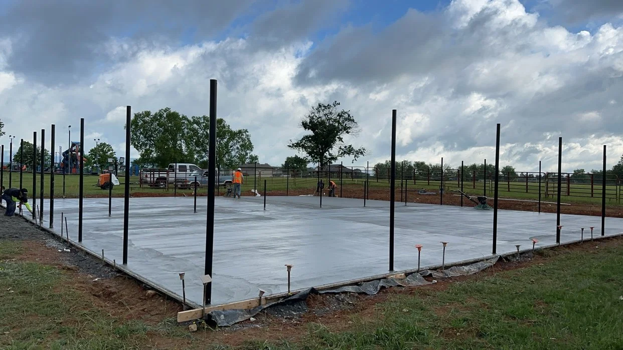 Construction site with freshly poured concrete foundation and steel posts for a building, under a cloudy sky.