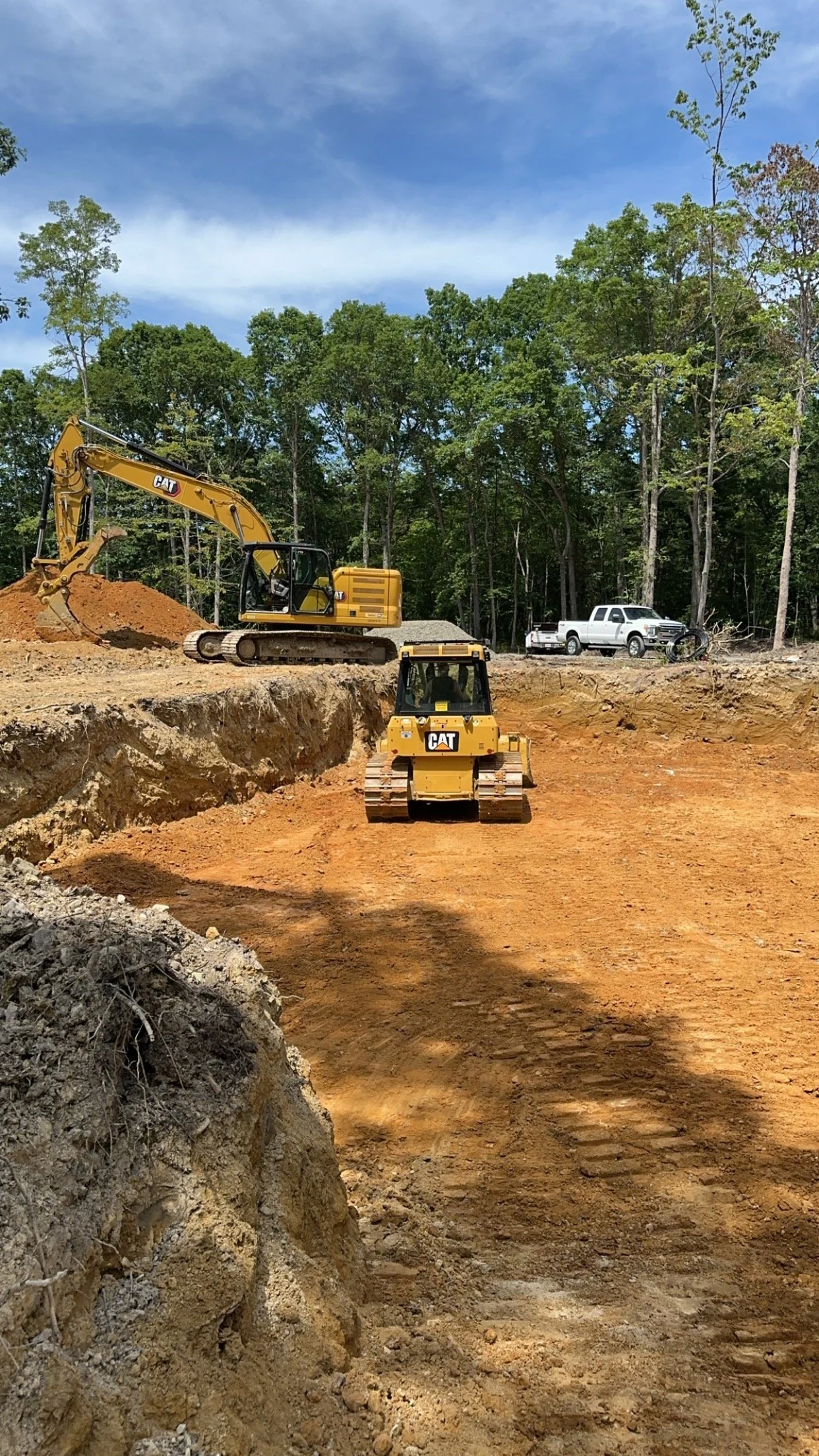 Construction site with two yellow Caterpillar bulldozers working on a dirt area, surrounded by trees and a blue sky.