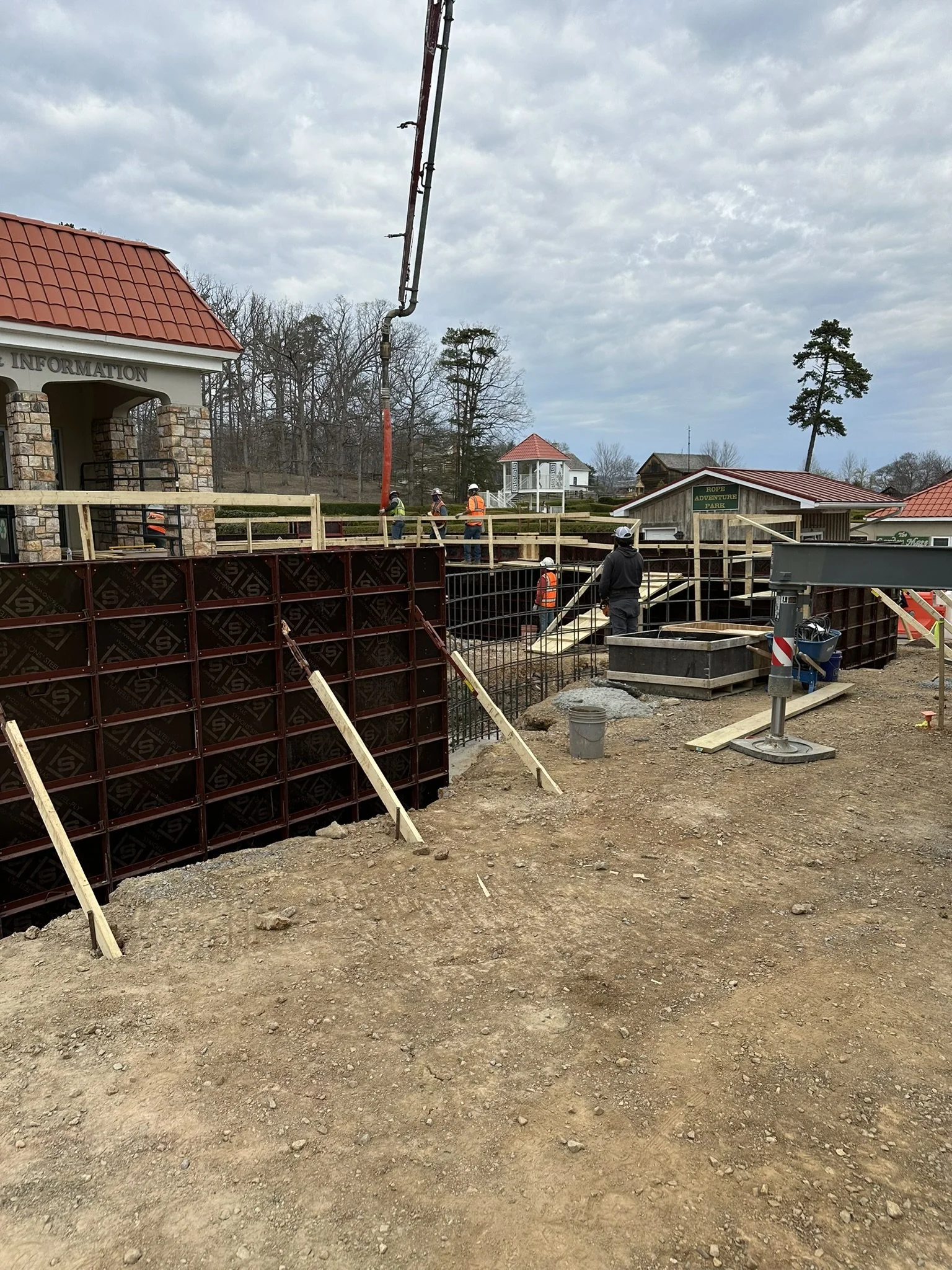 Construction site with workers installing a railing at an outdoor park or recreational area, with a small white pavilion and trees in the background.
