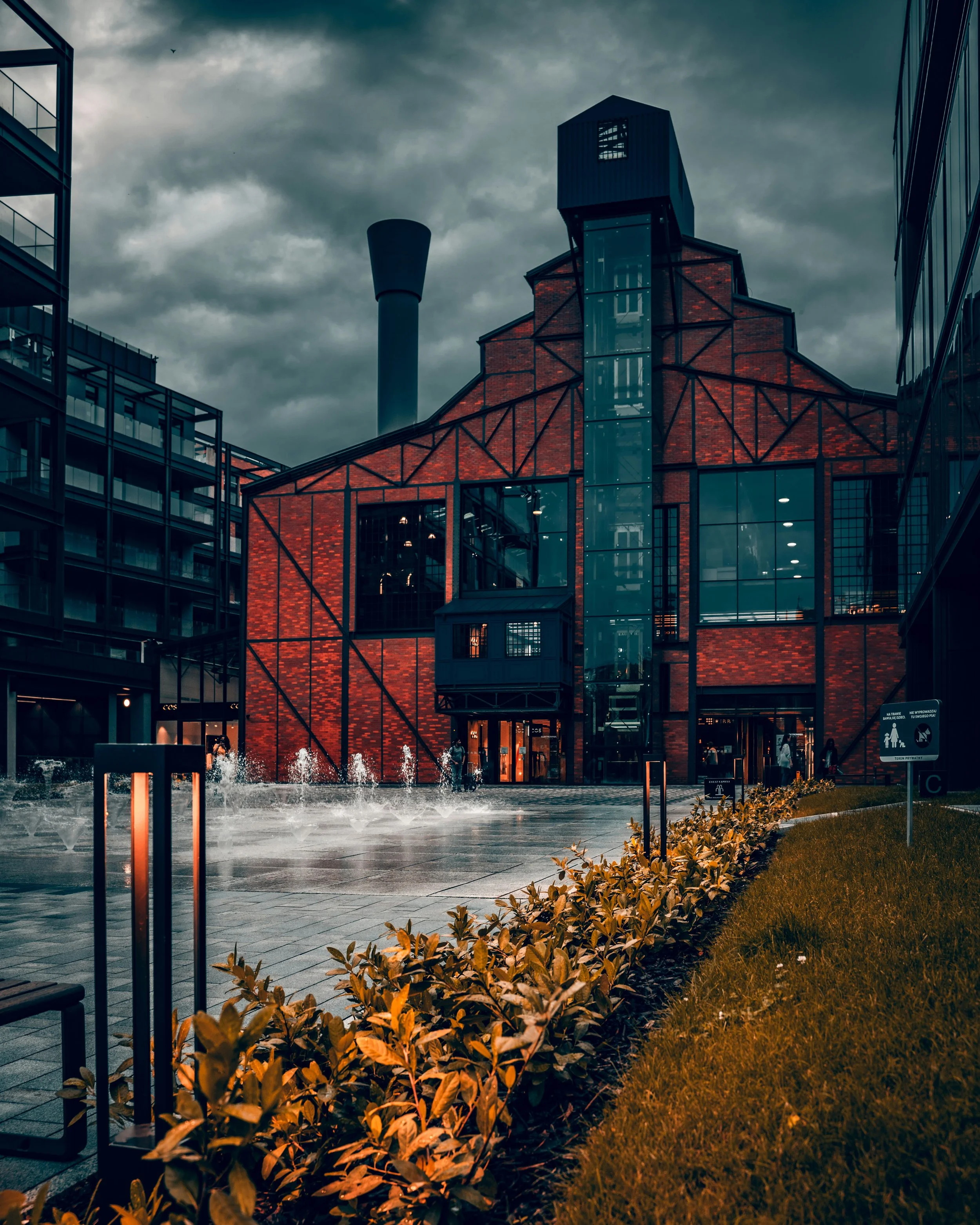 Urban scene featuring a modern red brick building with large glass windows, surrounded by other buildings, plants, and a fountain with water jets, under a cloudy sky.