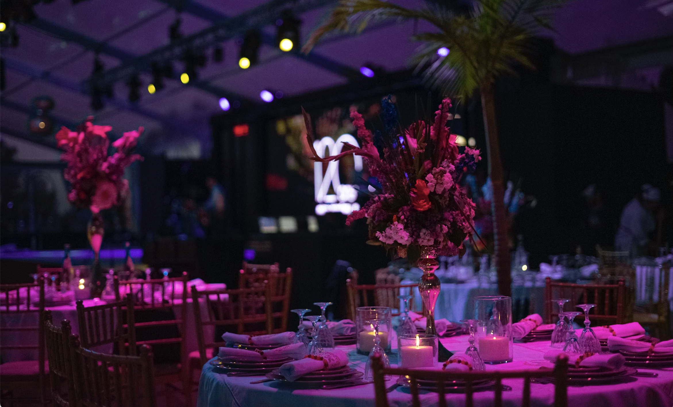 Elegantly decorated event venue with round tables, floral centerpieces, candles, and purple ambient lighting, set up for a celebration or banquet.