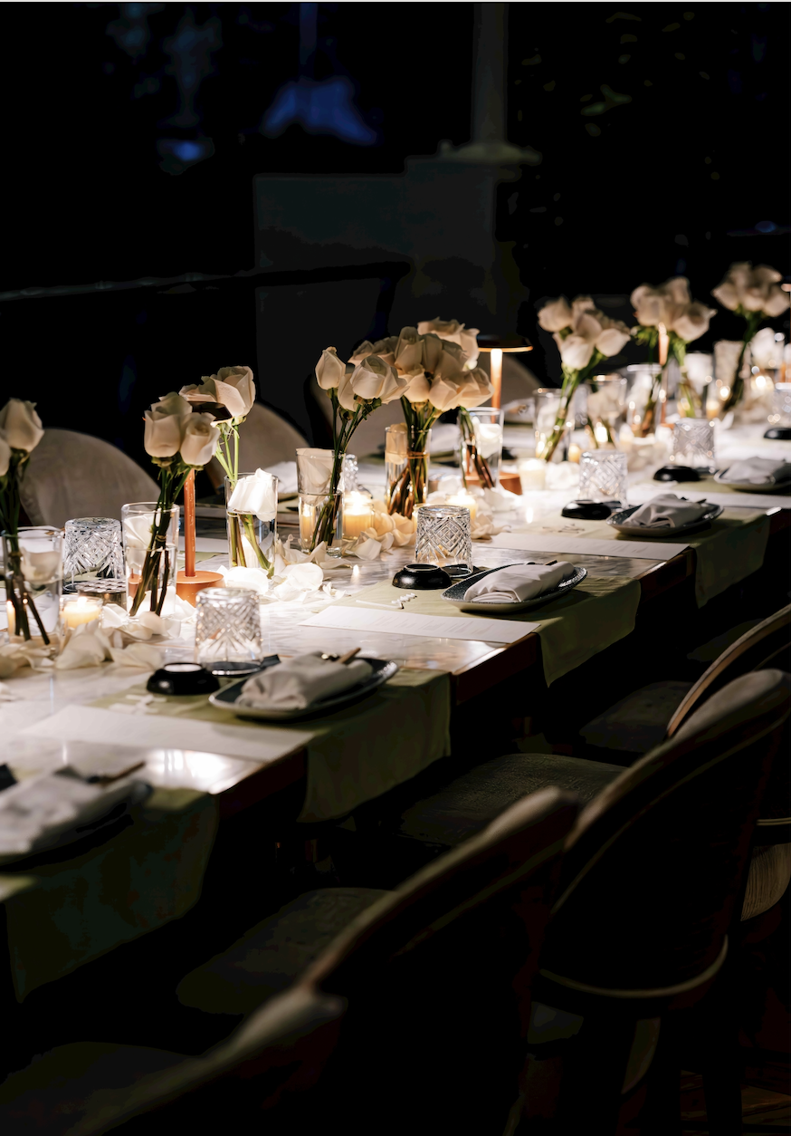 Elegant dining table set with white roses in glass vases, lit candles, wine glasses, and napkins in a dimly lit room, suitable for a formal event or wedding reception.