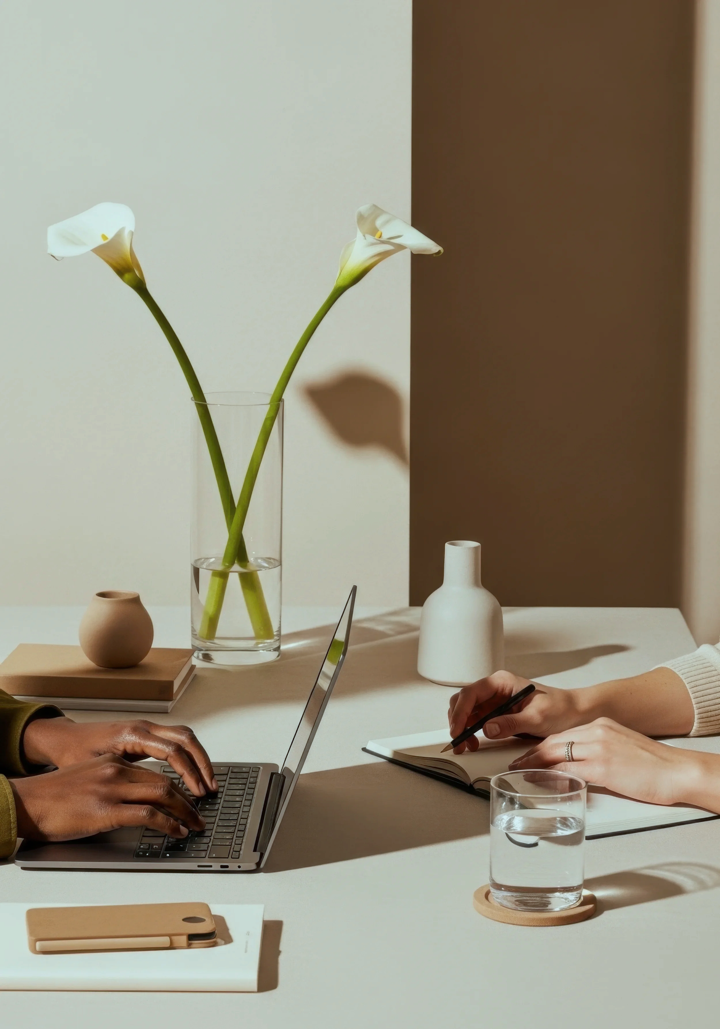 Two people working at a table with a laptop, notebook, pen, glass of water, and decorative vases, with a tall glass vase containing white flowers, in a modern, minimalistic setting.