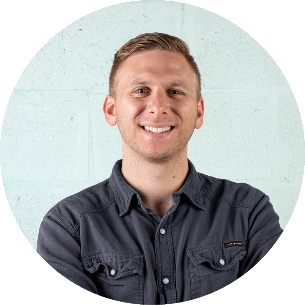 A man with short blond hair smiling and wearing a dark button-up shirt, standing in front of a light-colored wall.