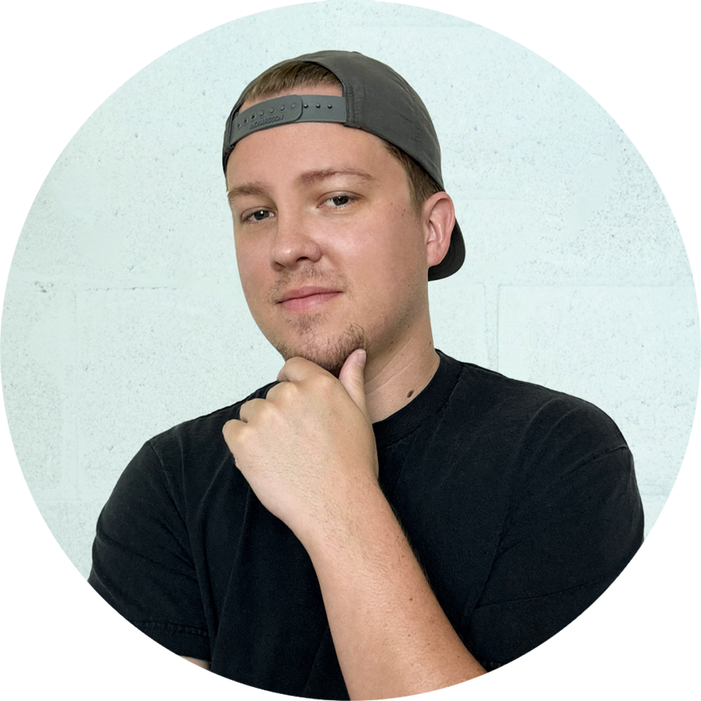 Young man with short hair wearing a backward gray cap and a black T-shirt, resting his chin on his hand in front of a light-colored wall.