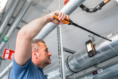 Un hombre trabaja en instalaciones industriales, sosteniendo una herramienta y observando un dispositivo móvil.