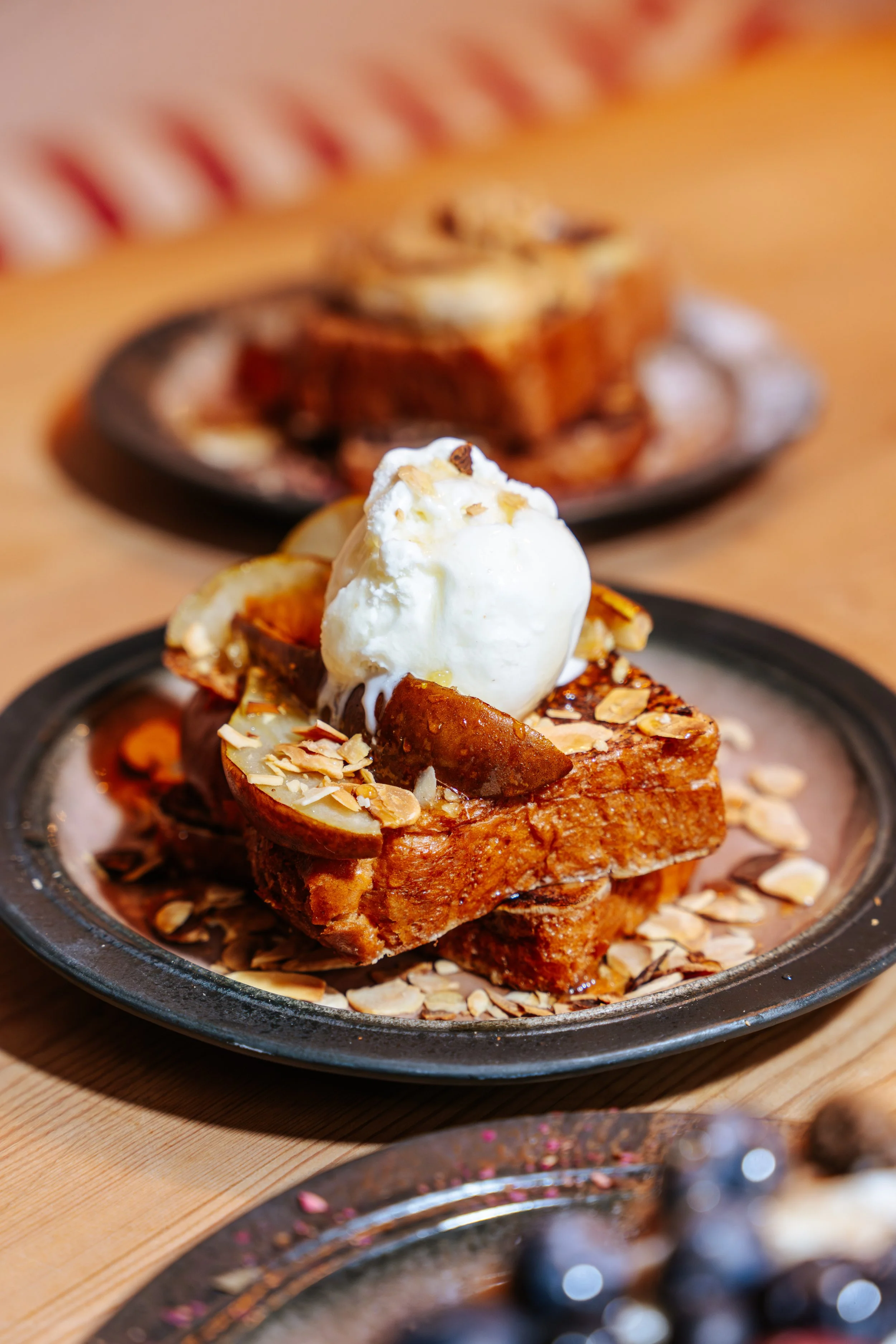 A plate of French toast topped with a scoop of vanilla ice cream, whipped cream, sliced almonds, and syrup, with another similar plate blurred in the background.