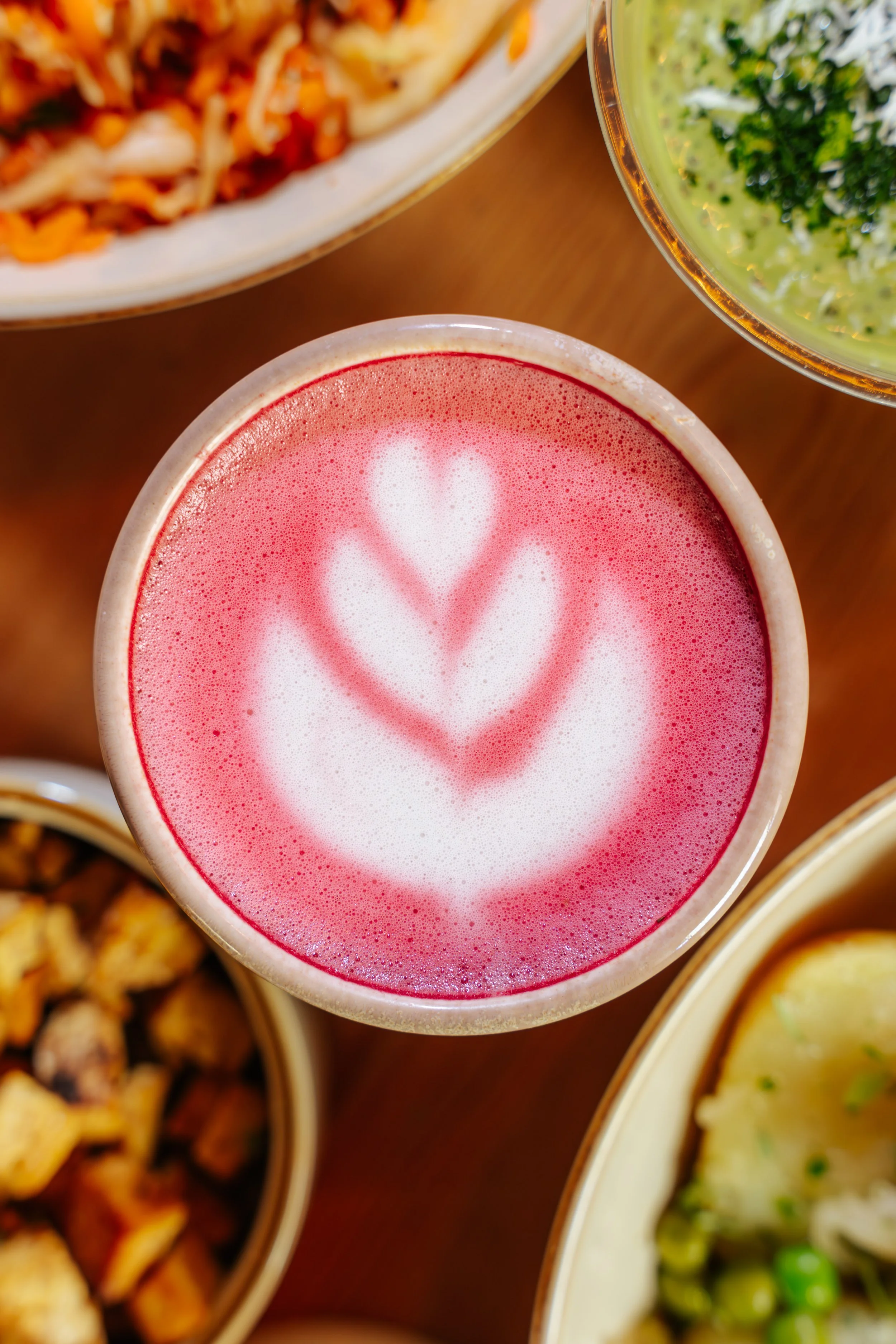 A pink beverage with a heart-shaped latte art design on top, surrounded by bowls of food on a wooden table.