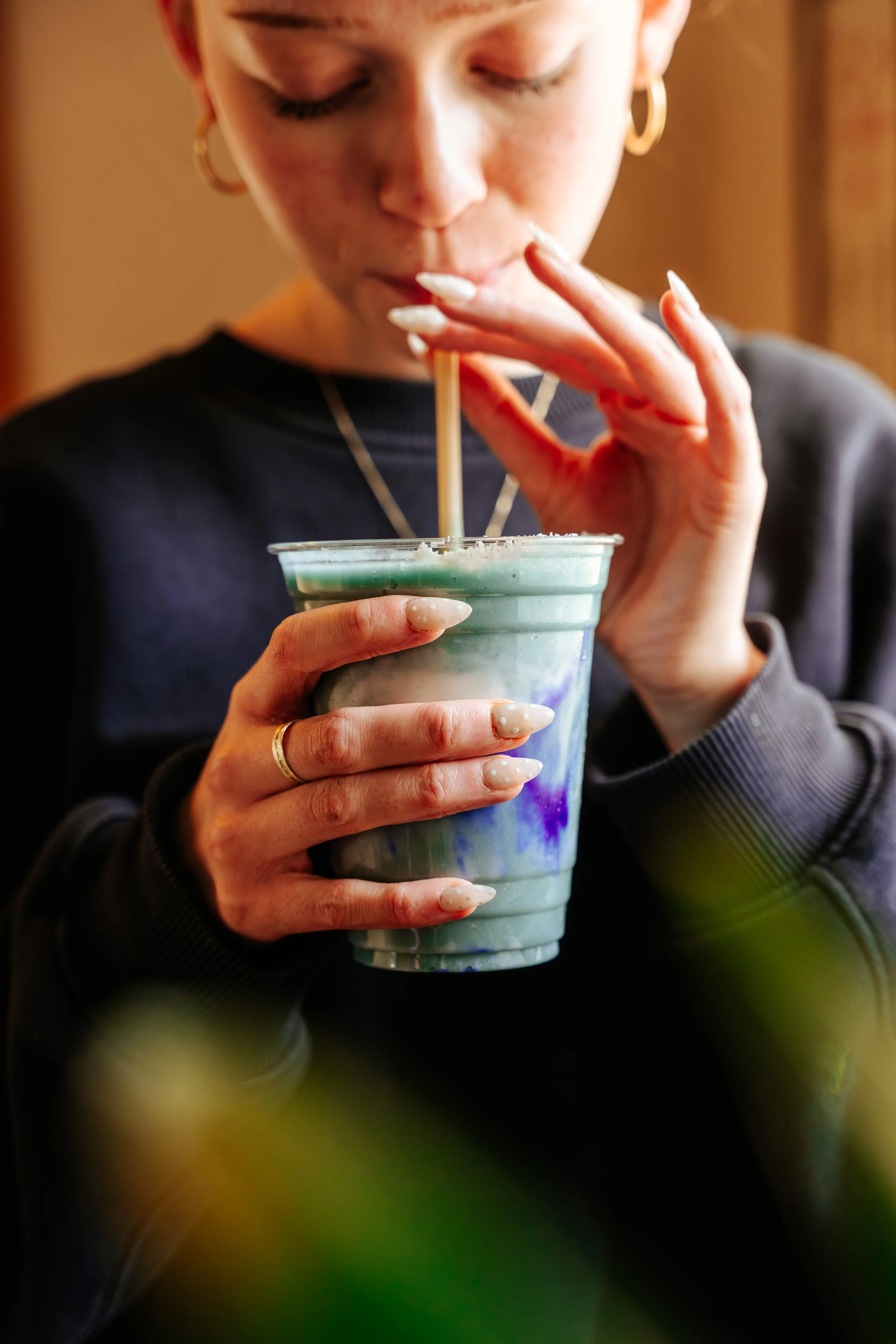 A woman with painted nails and gold jewelry drinking a colorful beverage through a straw from a clear plastic cup.