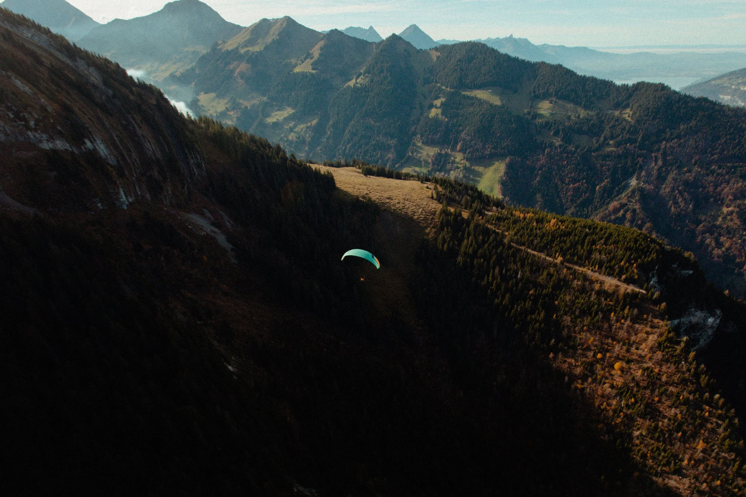 Two people paragliding over mountainous terrain with snow-capped peaks in the background. They are wearing helmets and sunglasses, with a bright sky and scenic valleys below.