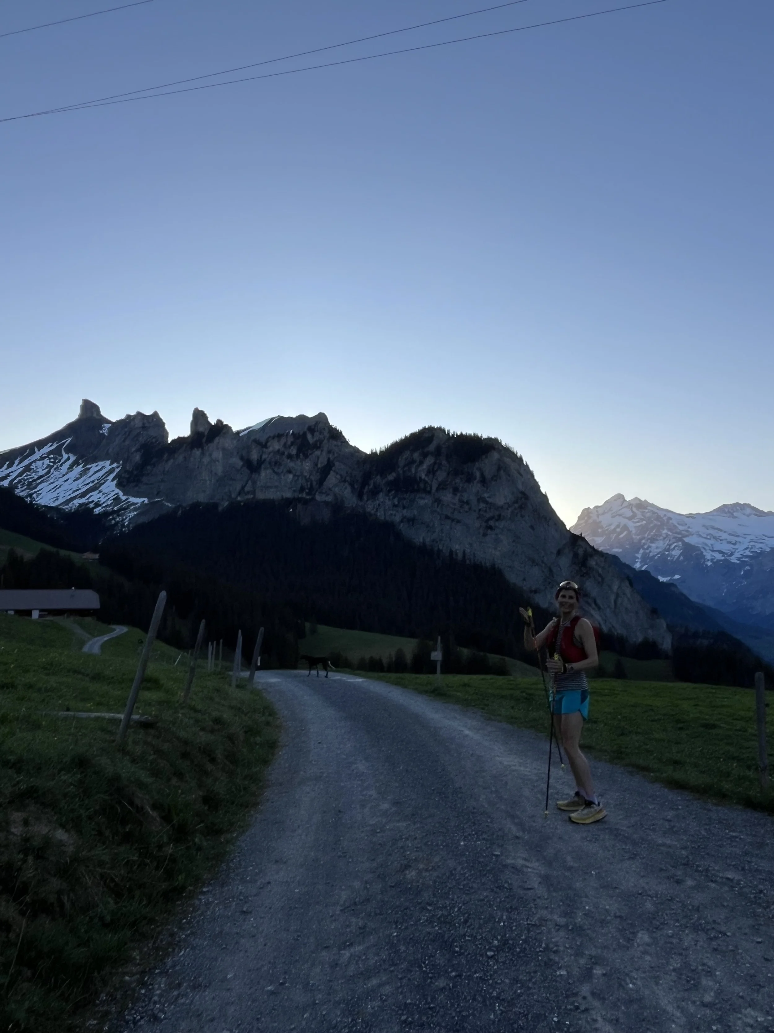 Silhouettes of five people standing on a mountain peak against a bright sun and clear sky, with distant mountains and contrails visible in the background.