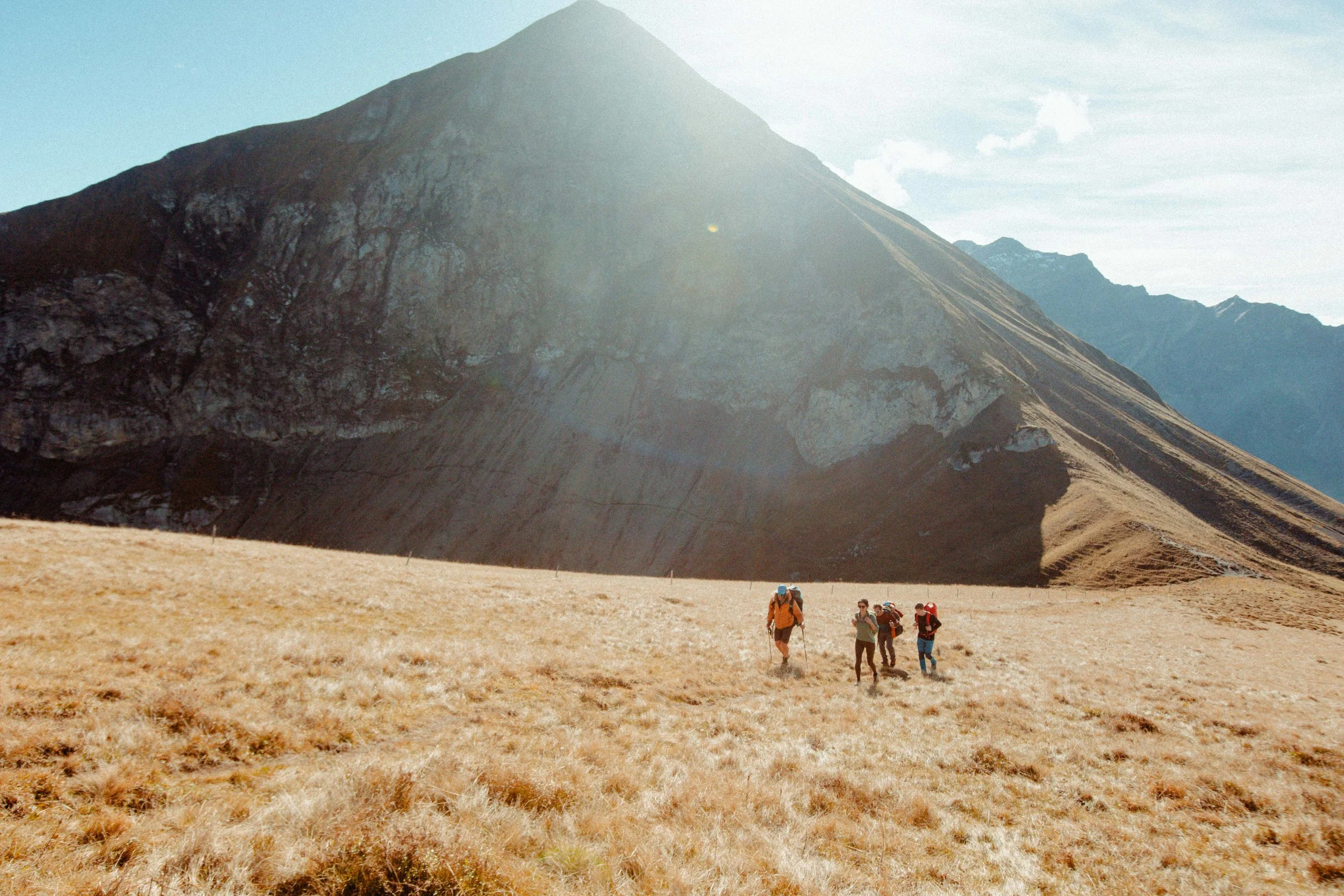 Two people paragliding over mountainous terrain with snow-capped peaks in the background. They are wearing helmets and sunglasses, with a bright sky and scenic valleys below.