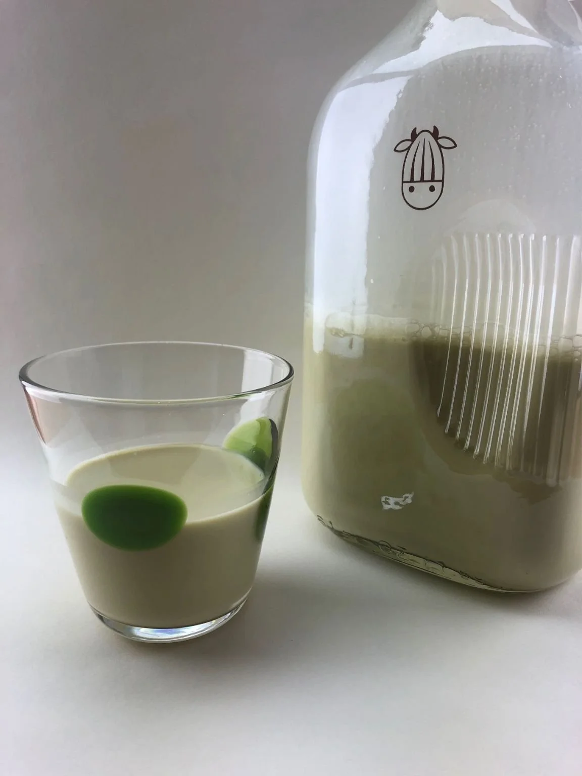 A glass of creamy beverage with green dots next to a large container of the same drink, which has a cow logo on it.