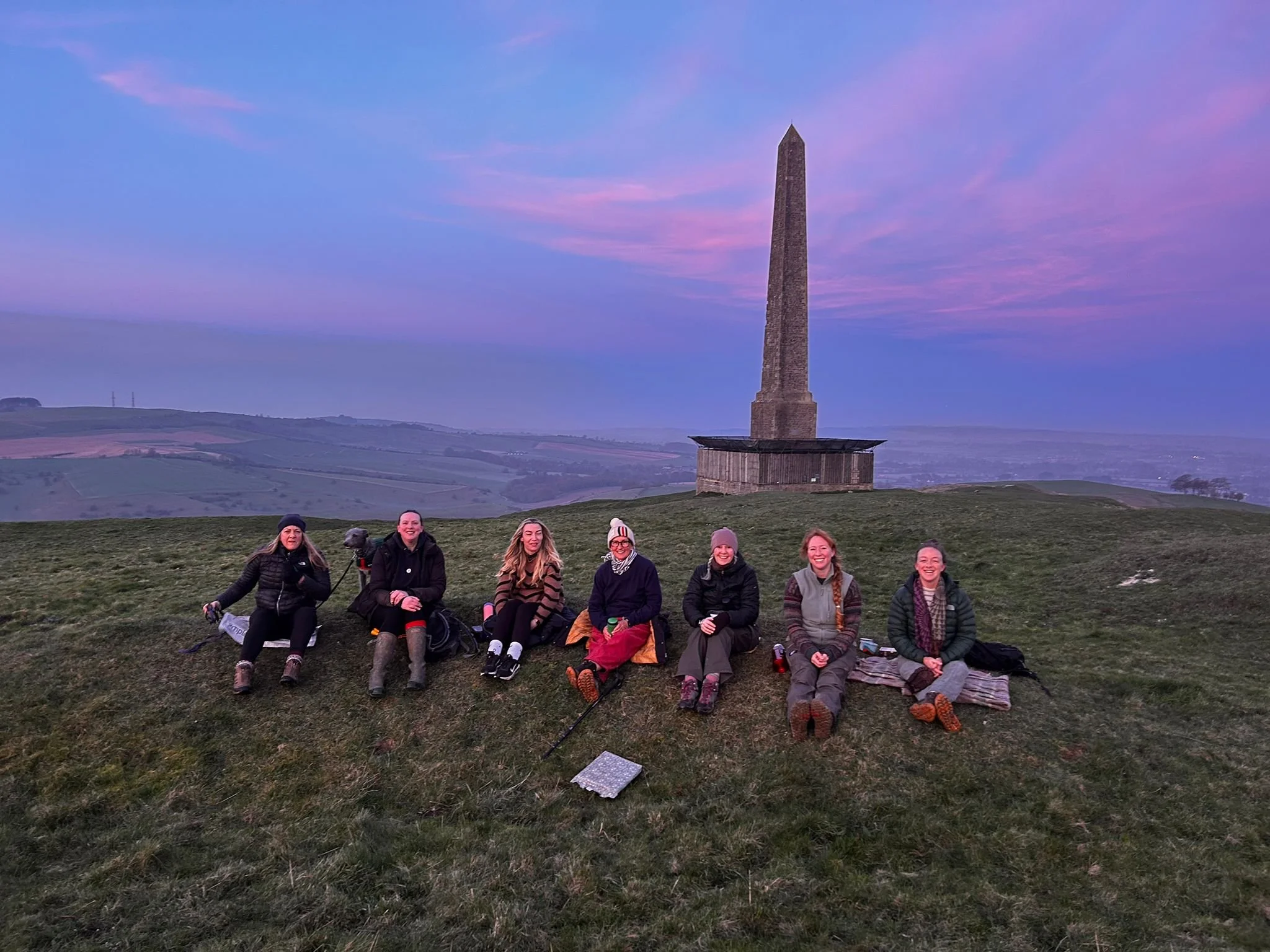 Group of seven women sitting on the grass with a memorial obelisk in the background during sunset.