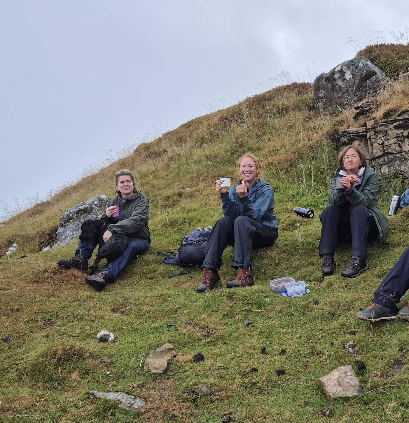 Three women sitting on a grassy hillside during a hike, drinking from cups, with rocks and a cloudy sky in the background.
