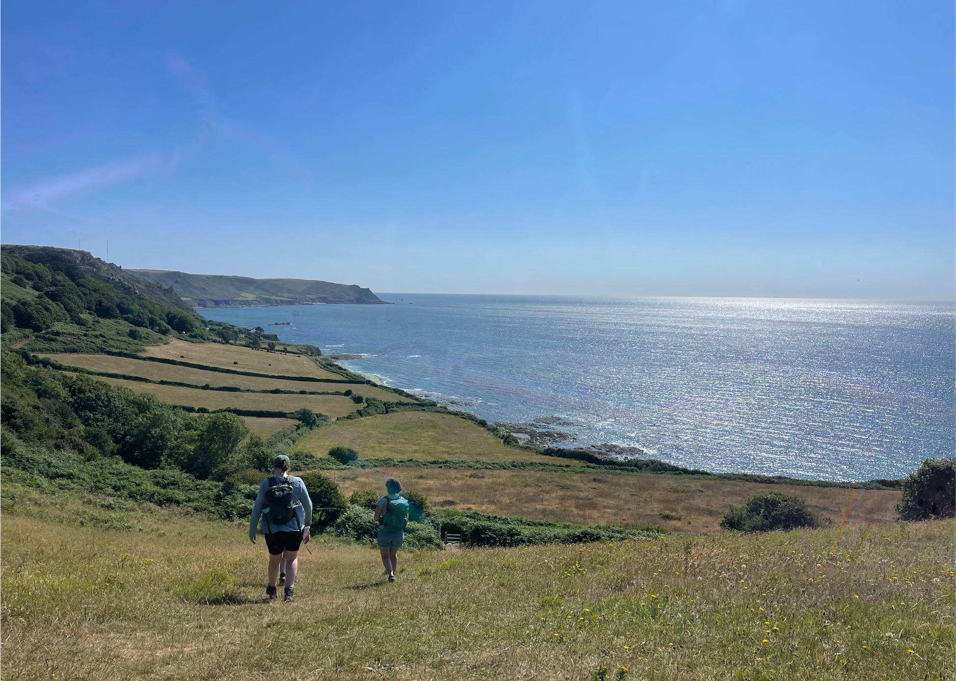 Two hikers walking down a grassy hill overlooking the ocean, with rolling green fields and cliffs in the background on a clear sunny day.
