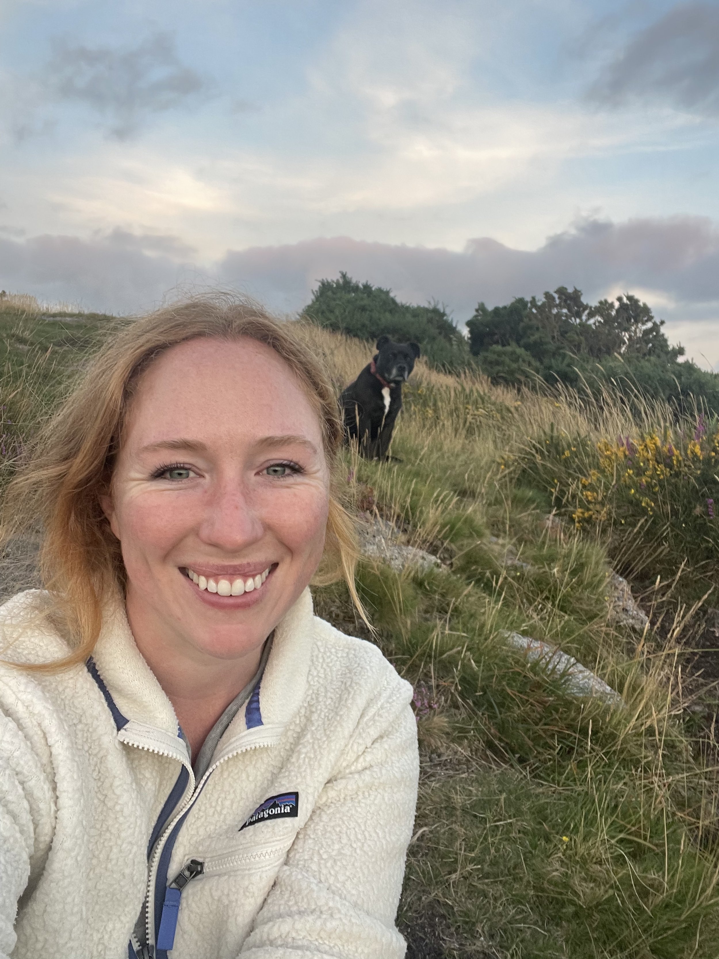 A woman with red hair smiling while taking a selfie outdoors on a grassy hillside with a black dog sitting behind her, under a cloudy sky.