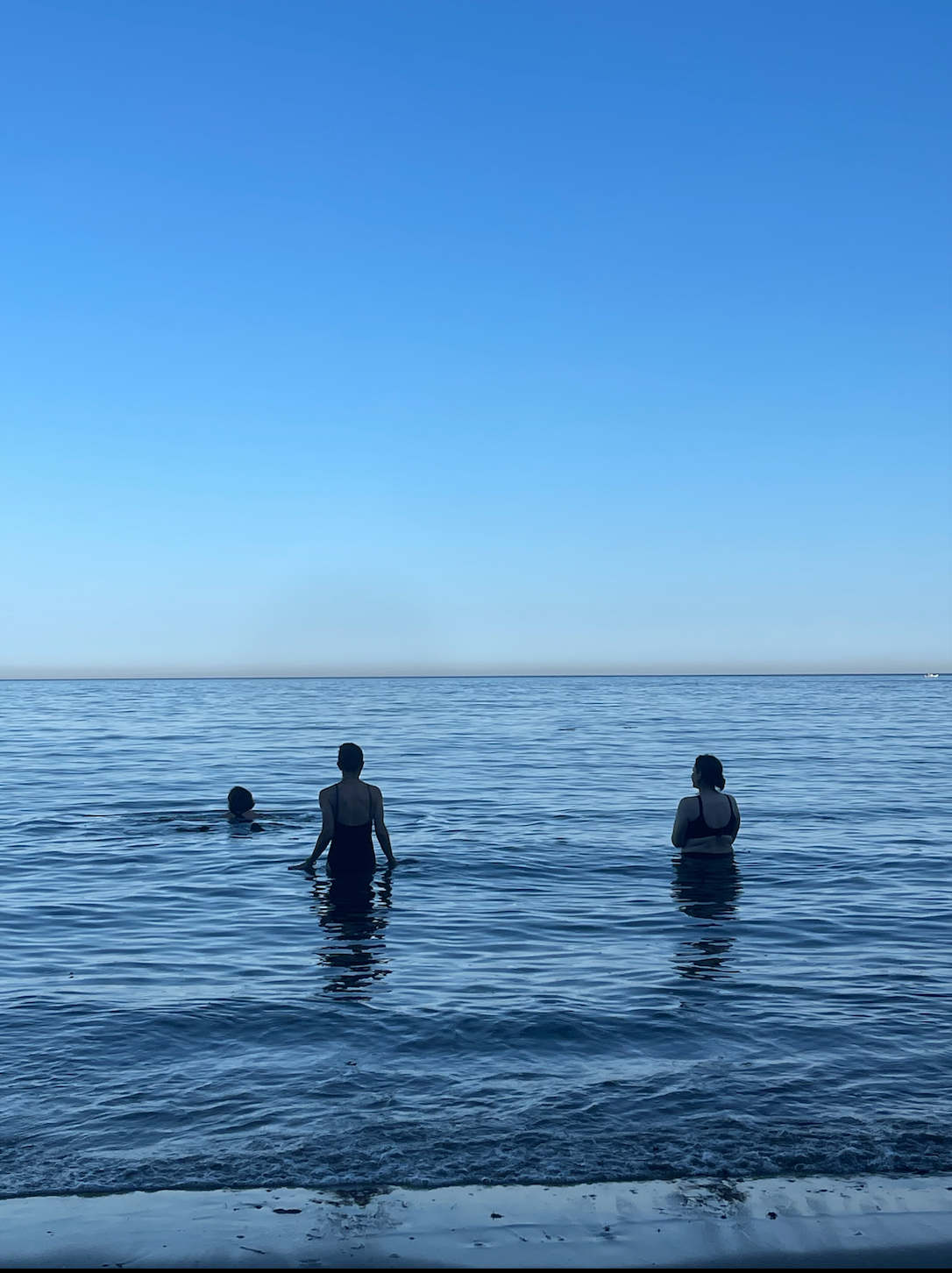 Three people standing in the ocean water at sunset or sunrise, with a clear blue sky above and calm waves.