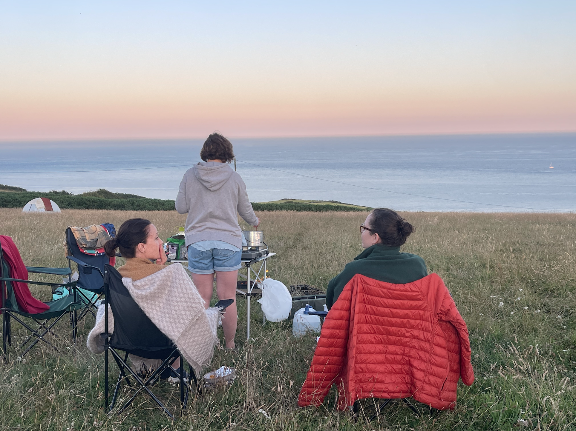 Three women sitting outdoors on camping chairs on a grassy hill, with a woman standing at a small table cooking, overlooking the ocean during sunset. East Prawle, South Devon
