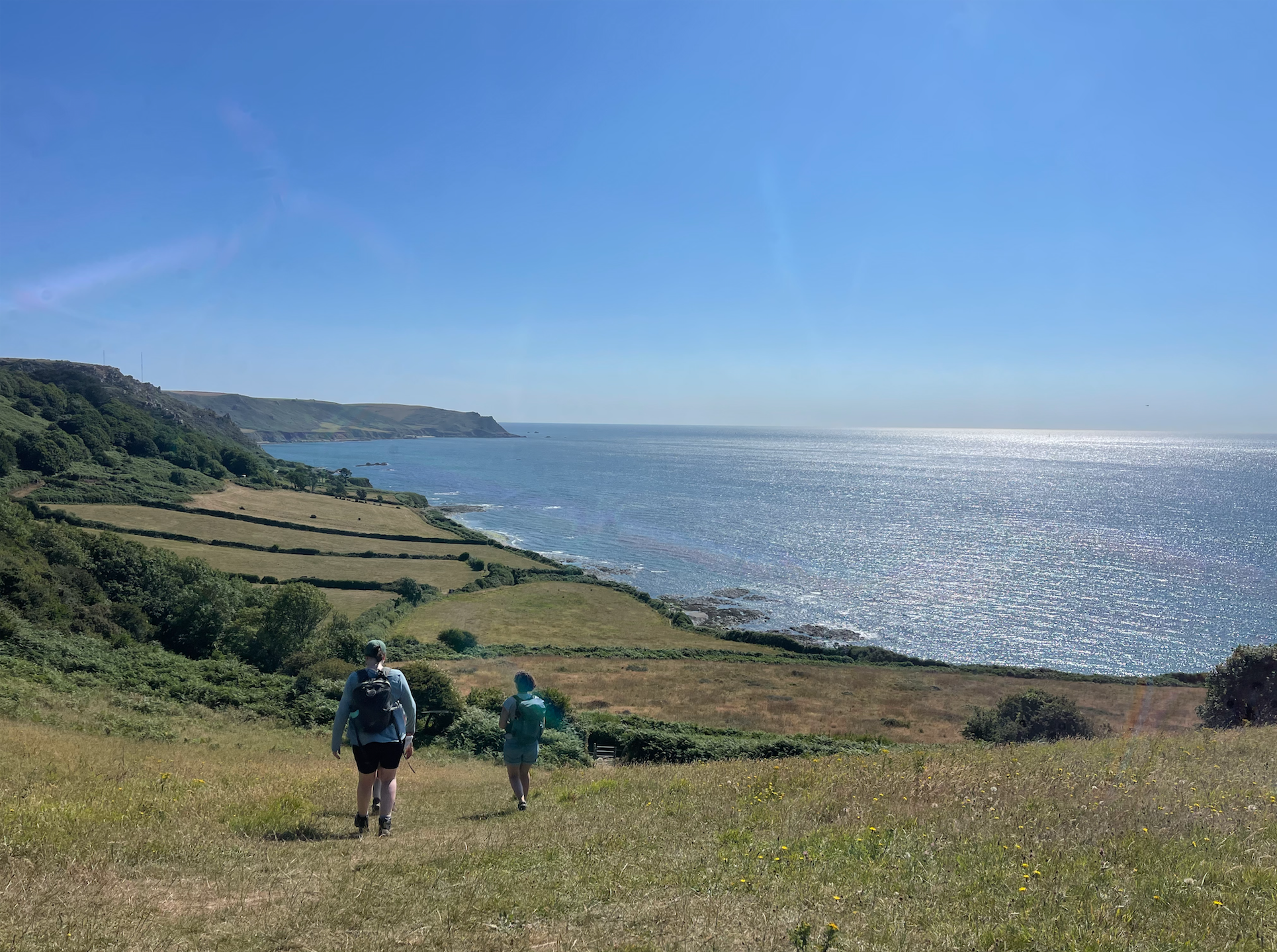 Two hikers walking on a grassy trail towards a coastline with cliffs, green fields, and the ocean under a clear blue sky. Prawle Point, South Devon